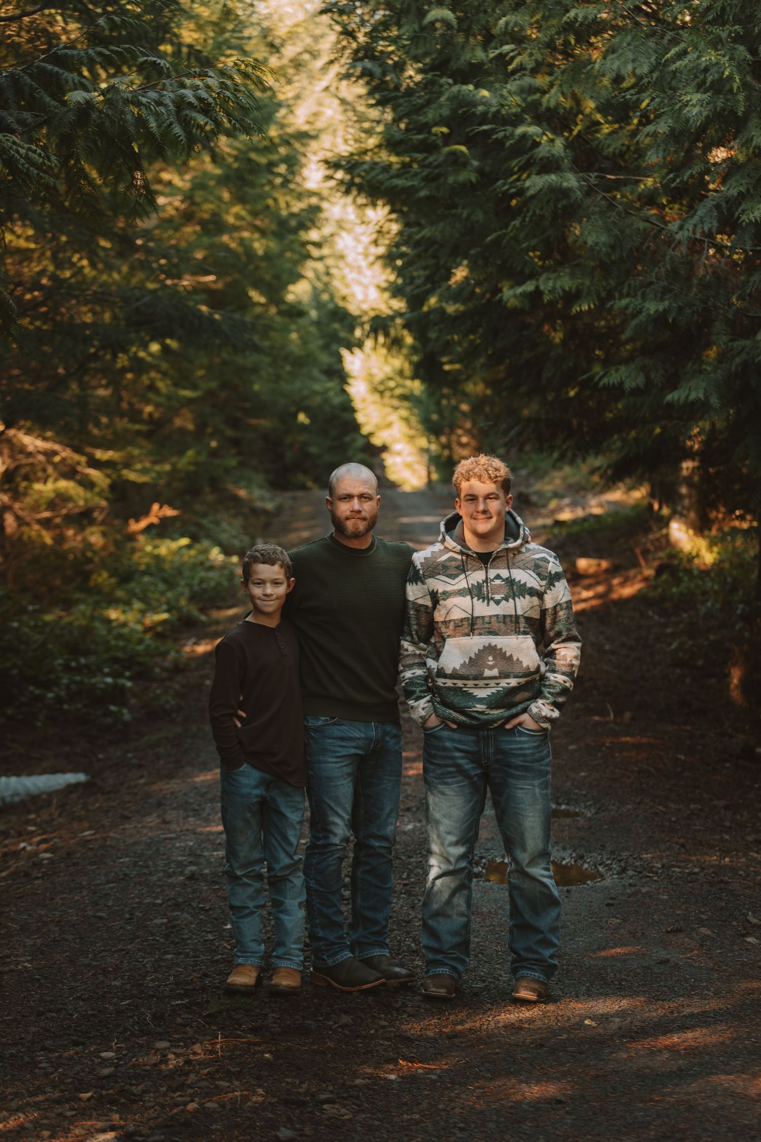 Three people stand together on a forest trail surrounded by evergreen trees at sunset.