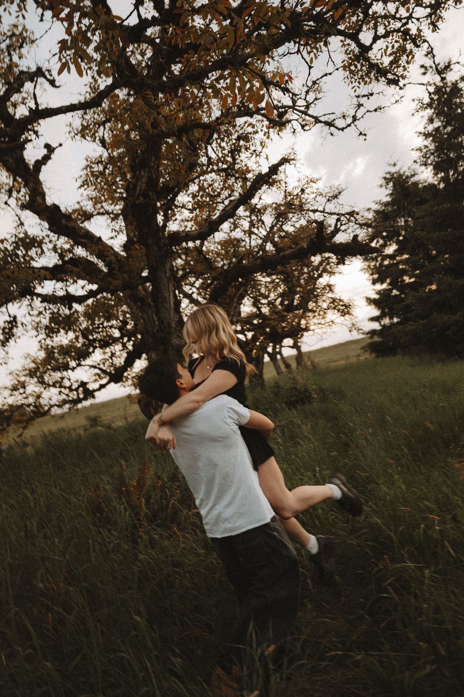 A man lifts and embraces a woman in a grassy field beside a large autumn tree at dusk.