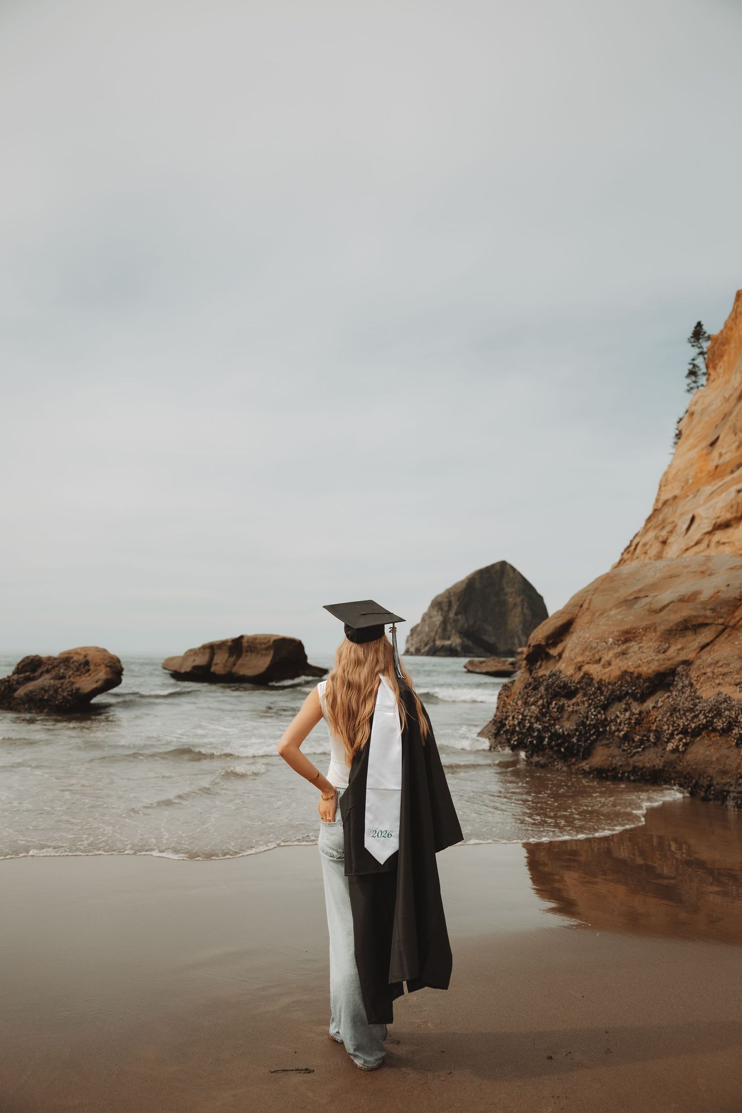 Graduate in cap and gown stands on a scenic beach with rocky outcrops and ocean waves on an overcast day.