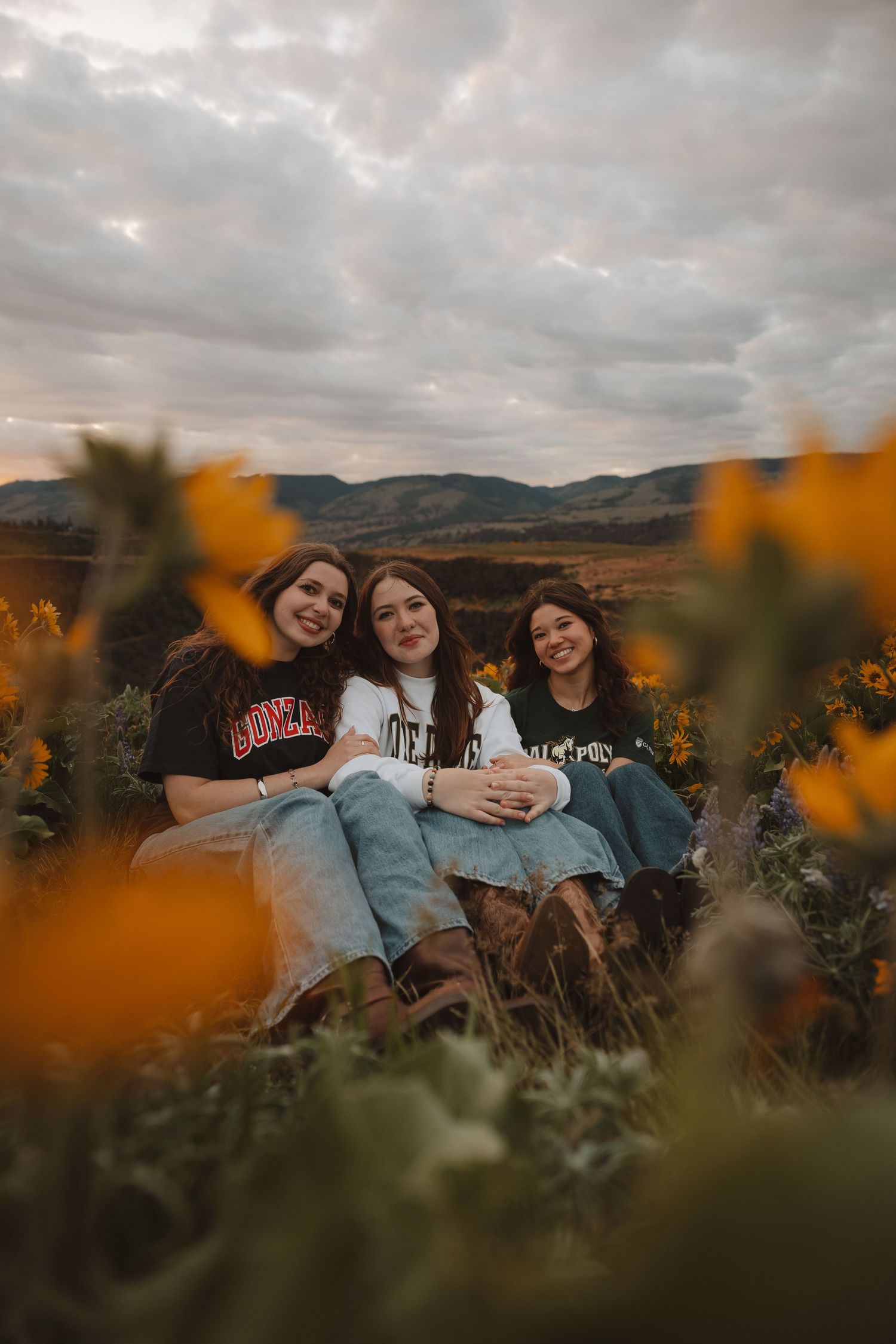 Three young women in university sweatshirts smile together while sitting in a field of yellow wildflowers at dusk.