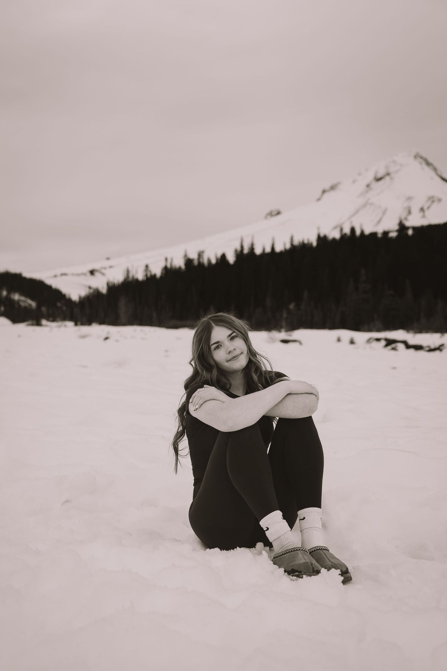 Young woman sitting in snow with mountains and pine trees in black and white winter landscape photo.