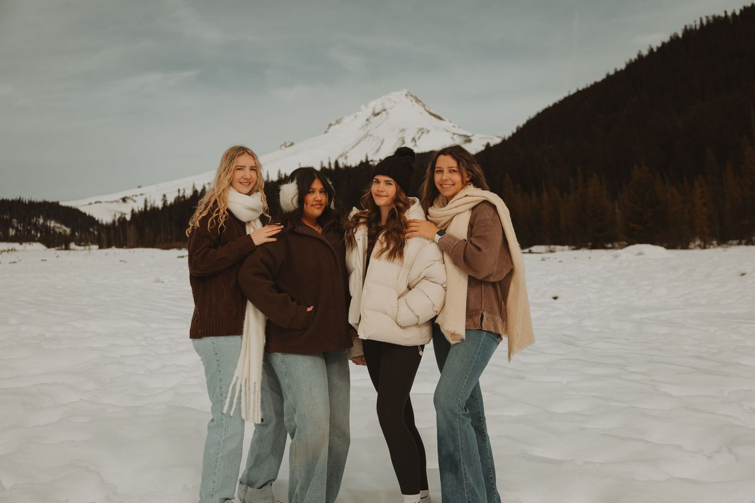 Four women pose together in snowy mountain landscape wearing winter coats and jeans with snow-capped peak behind.