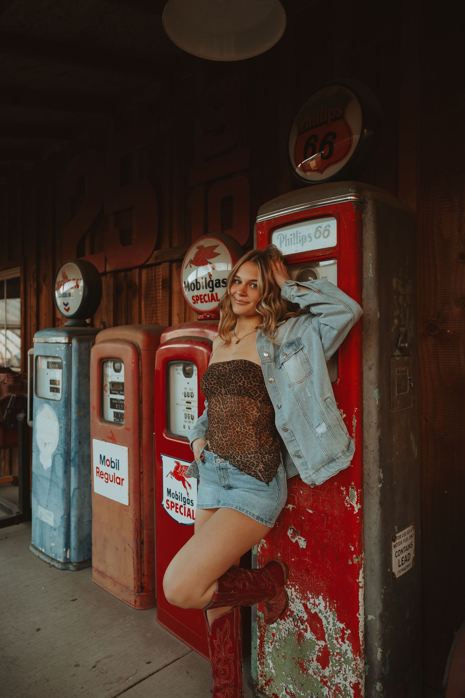 Young woman in denim jacket and jean shorts posing beside vintage Phillips 66 gas pumps at retro service station.