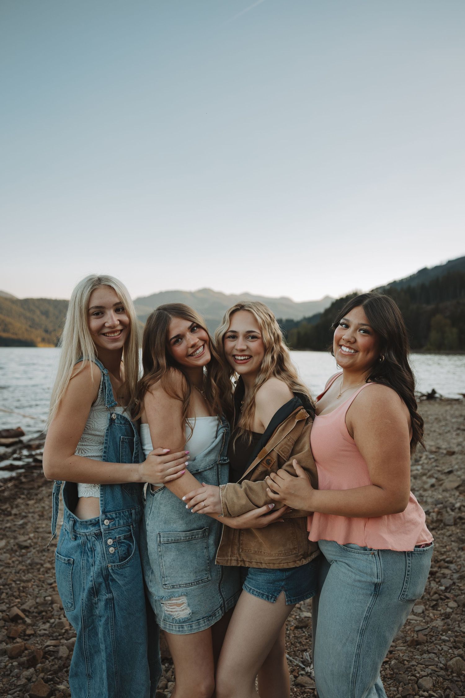 Four young women smiling and embracing on a rocky lakeshore with mountains in the background.
