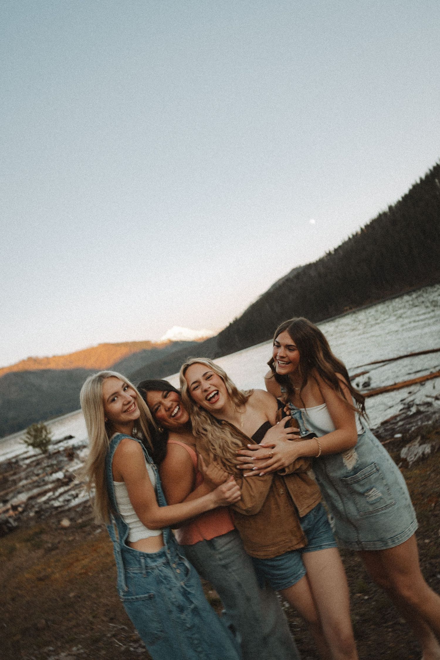 Four laughing women embrace on a mountain lakeshore during a vibrant sunset.