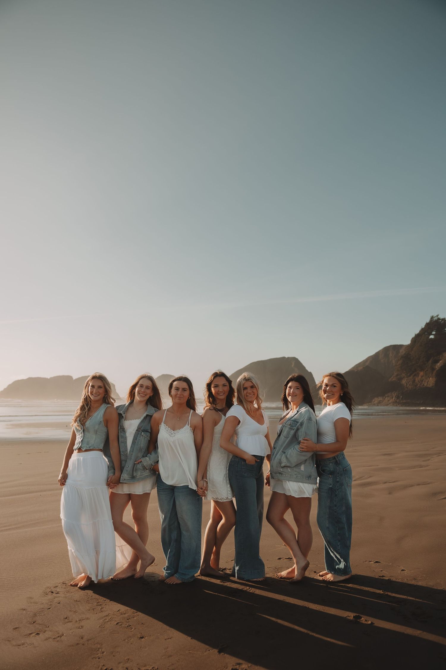 Seven women pose together on a sandy beach at golden hour wearing white and denim outfits.