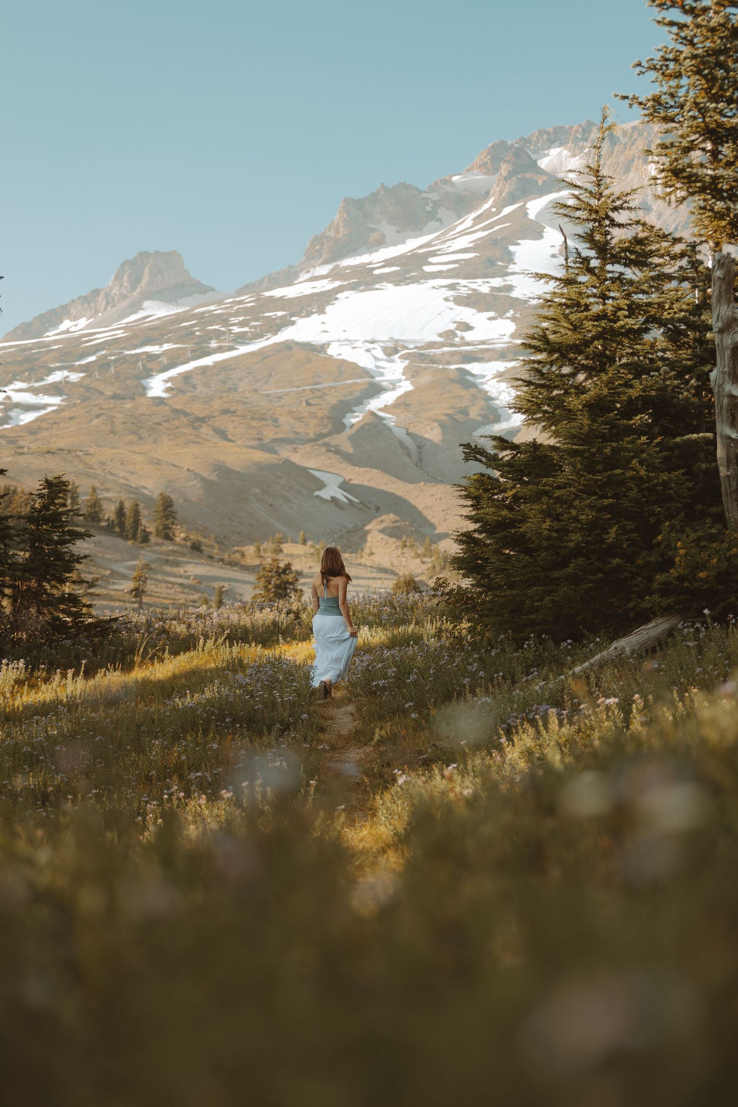 Woman in white dress walking through alpine meadow with snow-capped mountain peaks and evergreen trees in background.