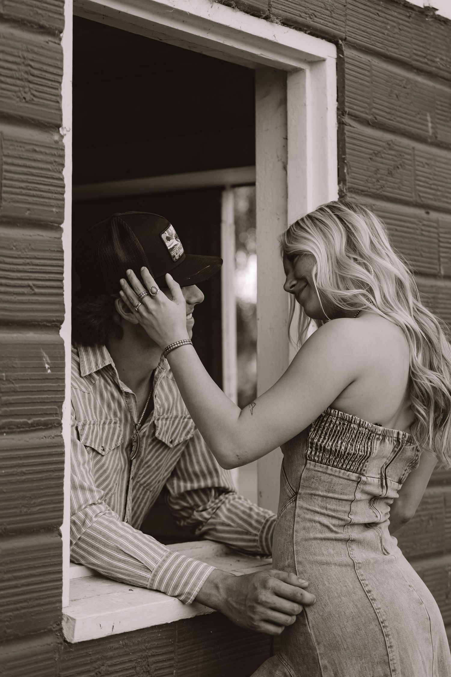 Black and white photo of a smiling blonde woman adjusting a mans cap through a barn window.