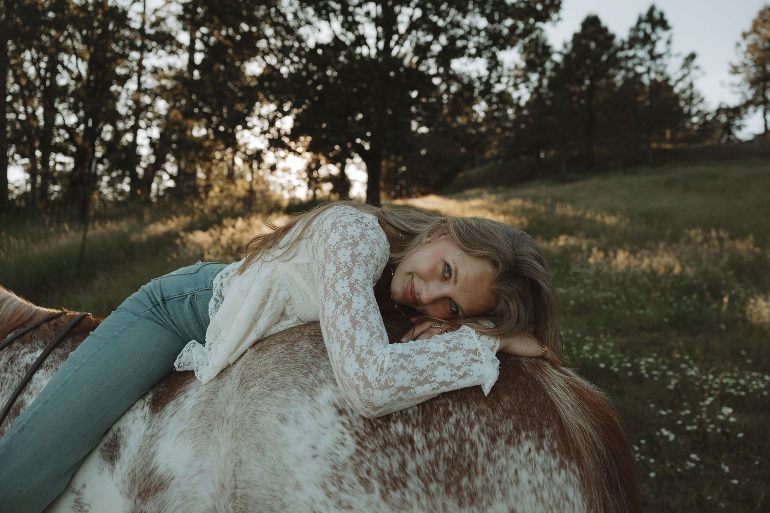Young girl laying on a horse in a sunlit meadow, wearing lace top and teal pants.