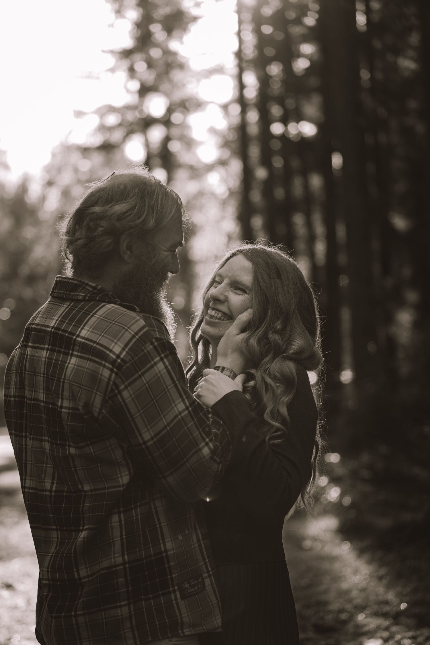 A couple in plaid clothing embraces and laughs together in a moody forest setting.