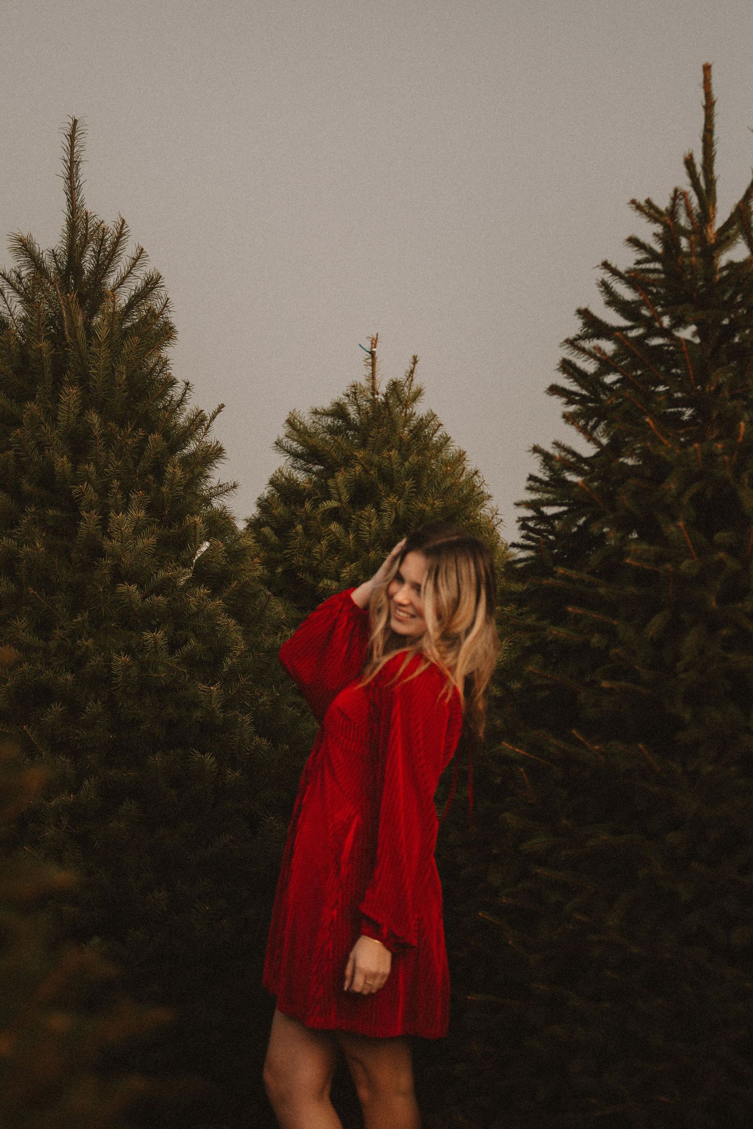 A person in a flowing red dress dances freely among evergreen trees at dusk.