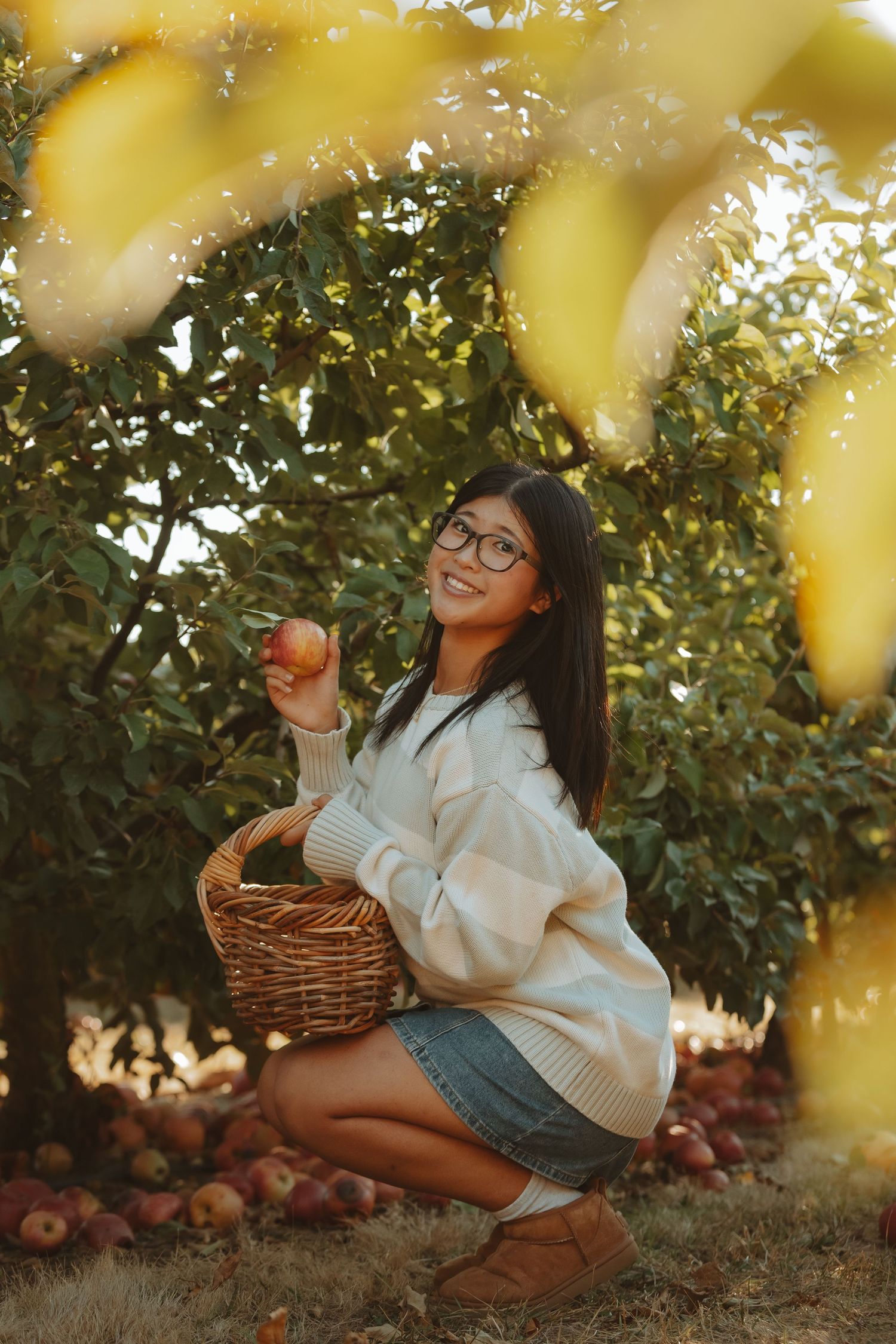 Someone in a cozy sweater sits near an apple tree with a woven basket during golden hour harvest time.