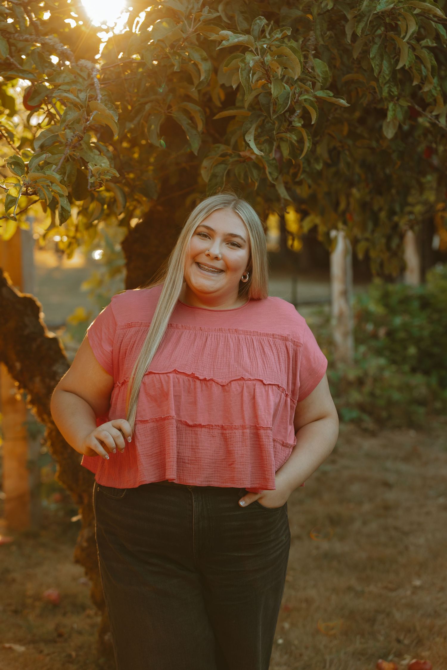 A person in a coral ruffled top and dark pants poses outdoors during golden hour among trees and foliage.