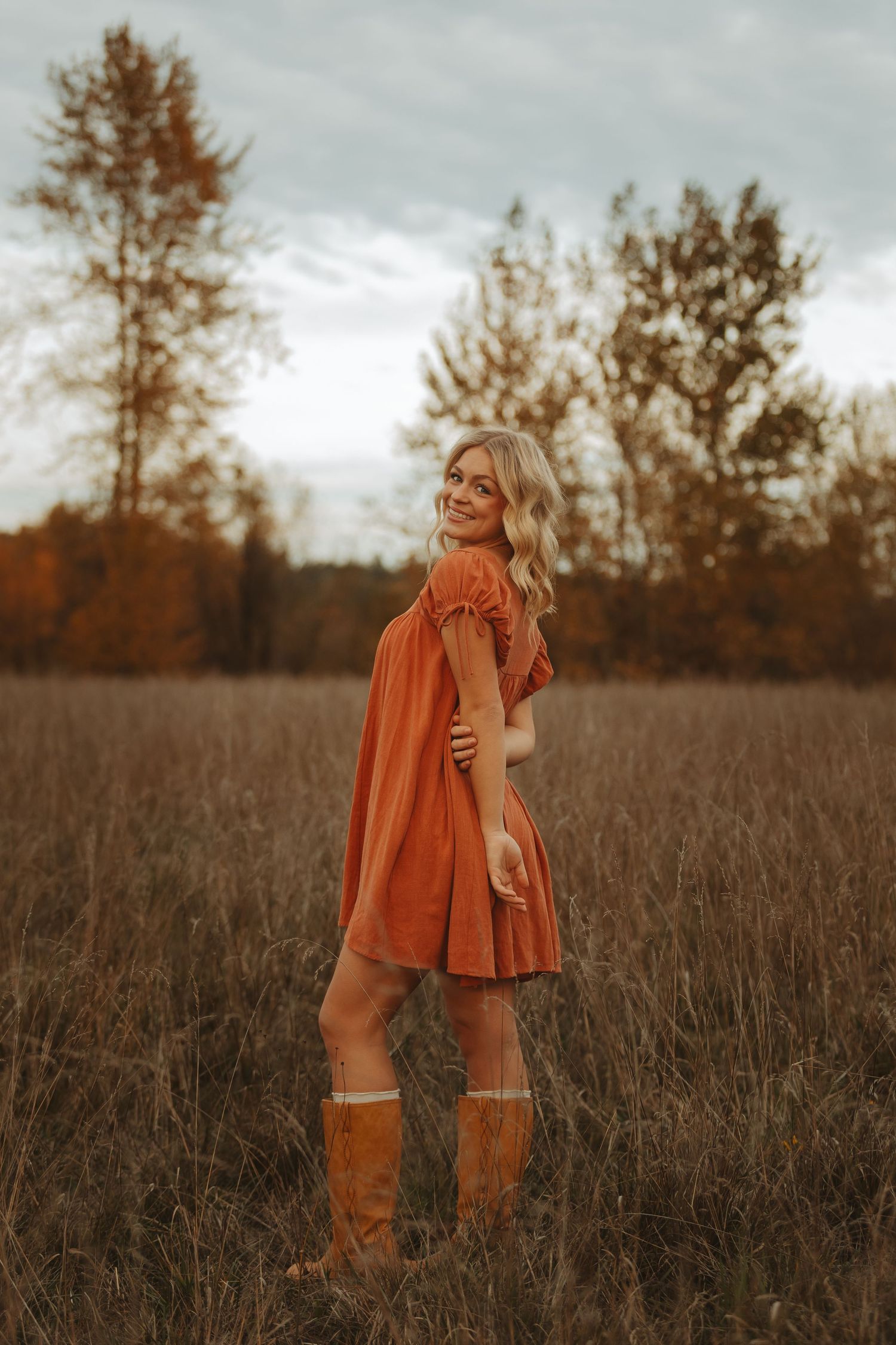 Person in rust-colored dress and knee-high boots stands among tall grass in autumn field.
