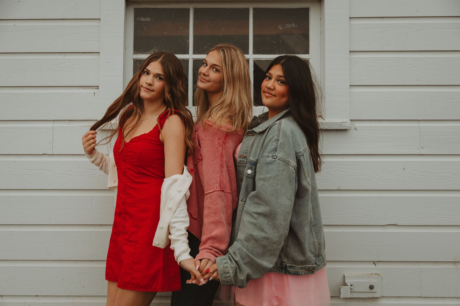 Three friends in casual dresses and jackets pose together against a white building exterior.