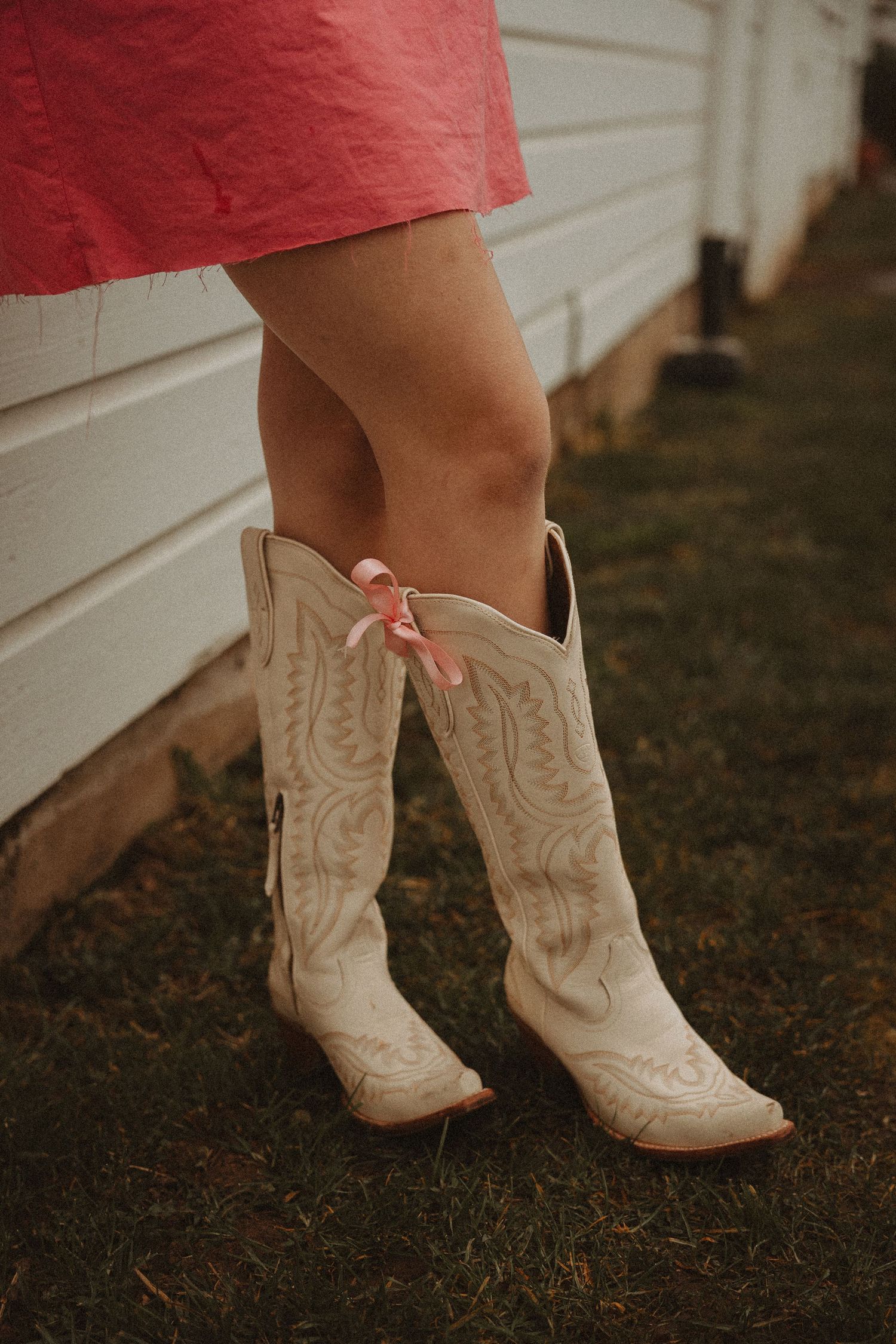 Close-up of white cowboy boots with embroidered details on grass against white building.