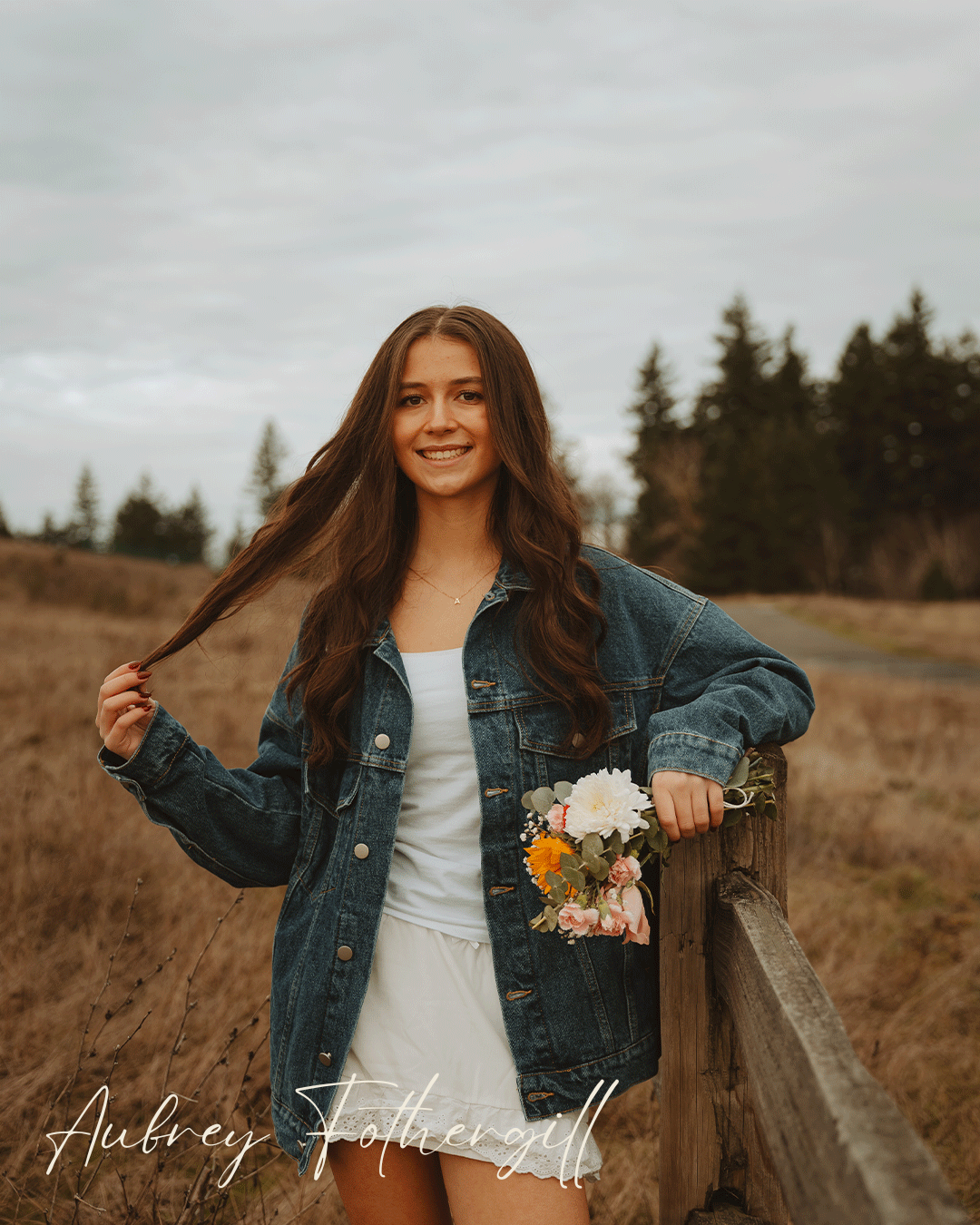 Moody outdoor portrait session in a golden field with denim jacket and white dress against overcast autumn sky.