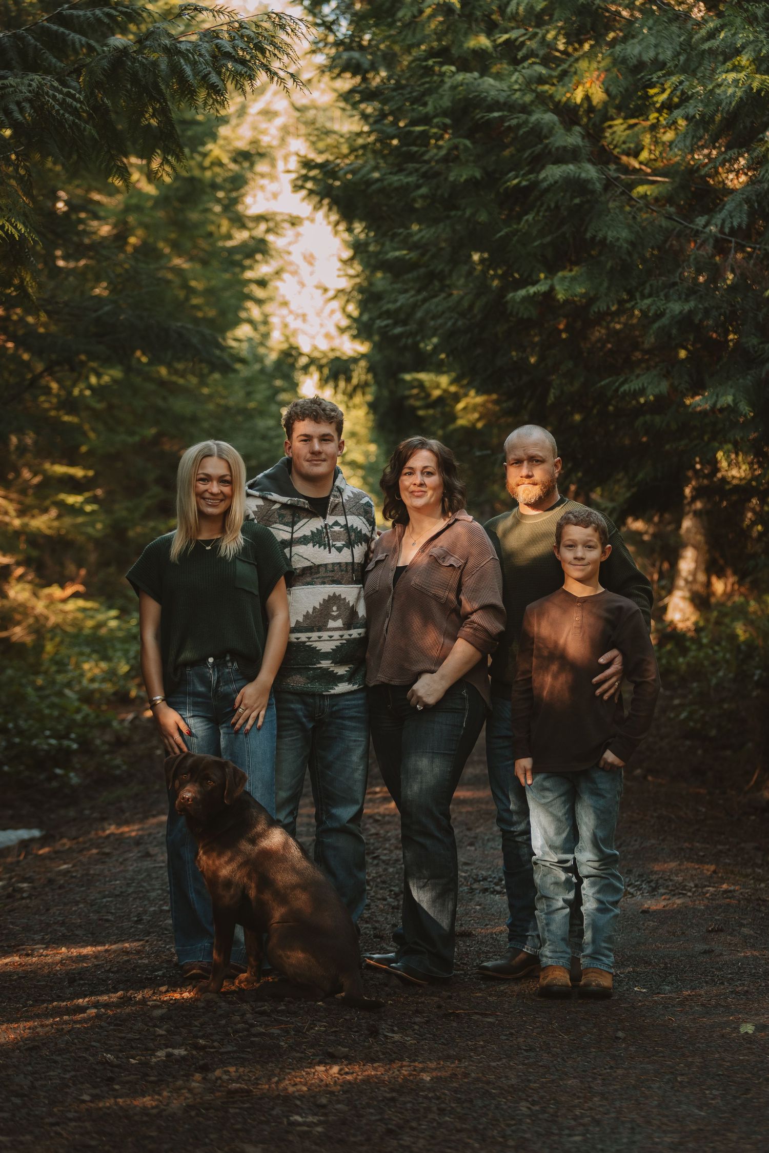 A family and their dog pose together in a wooded outdoor setting during golden hour lighting.