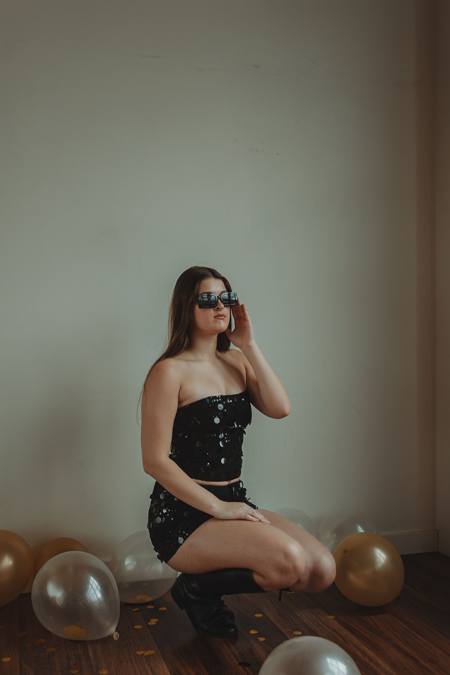 Woman in sequined black dress poses with silver balloons on wooden floor during photo shoot.