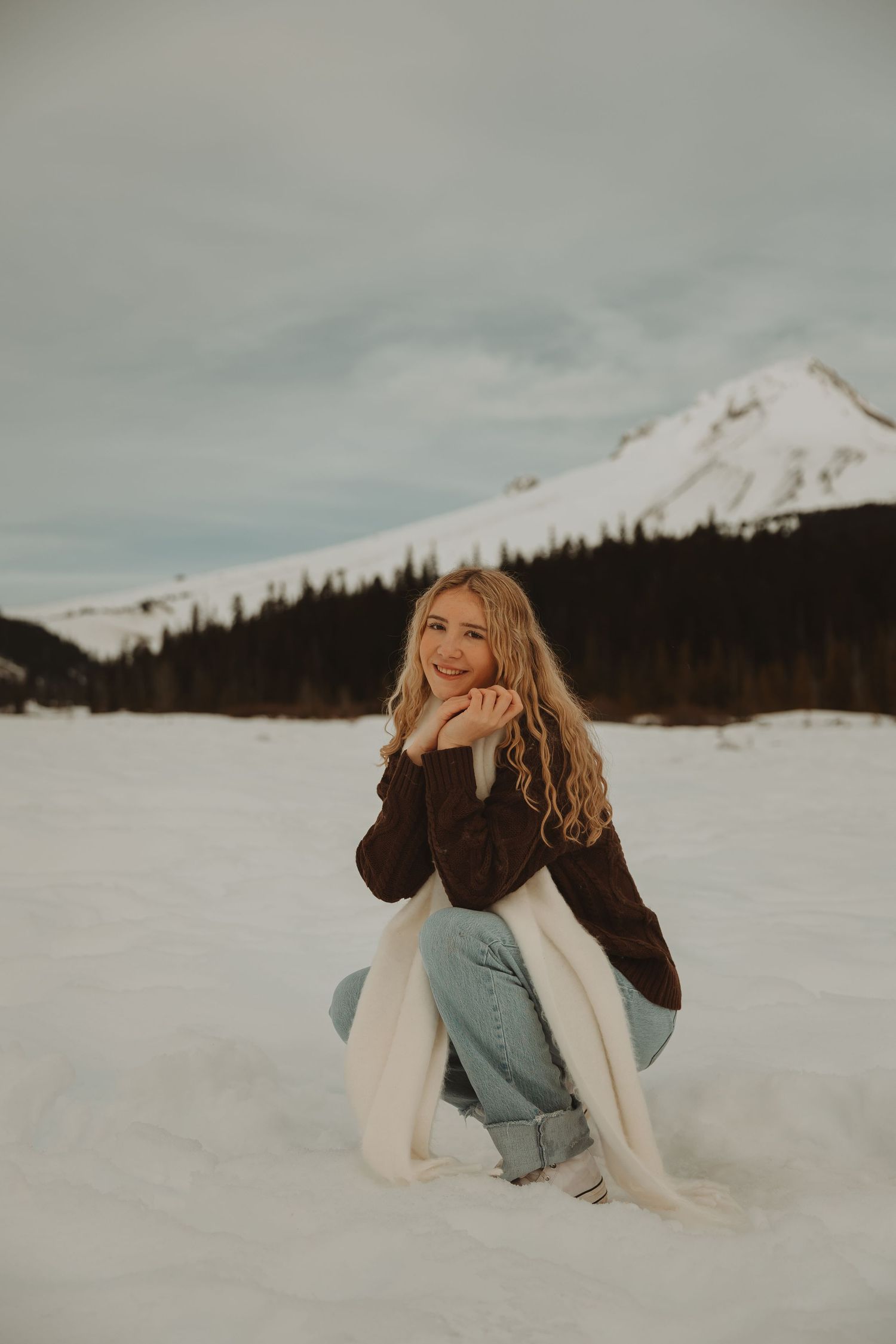 Smiling blonde woman crouching in snow wearing brown jacket, white scarf, and jeans with snowy mountain backdrop.