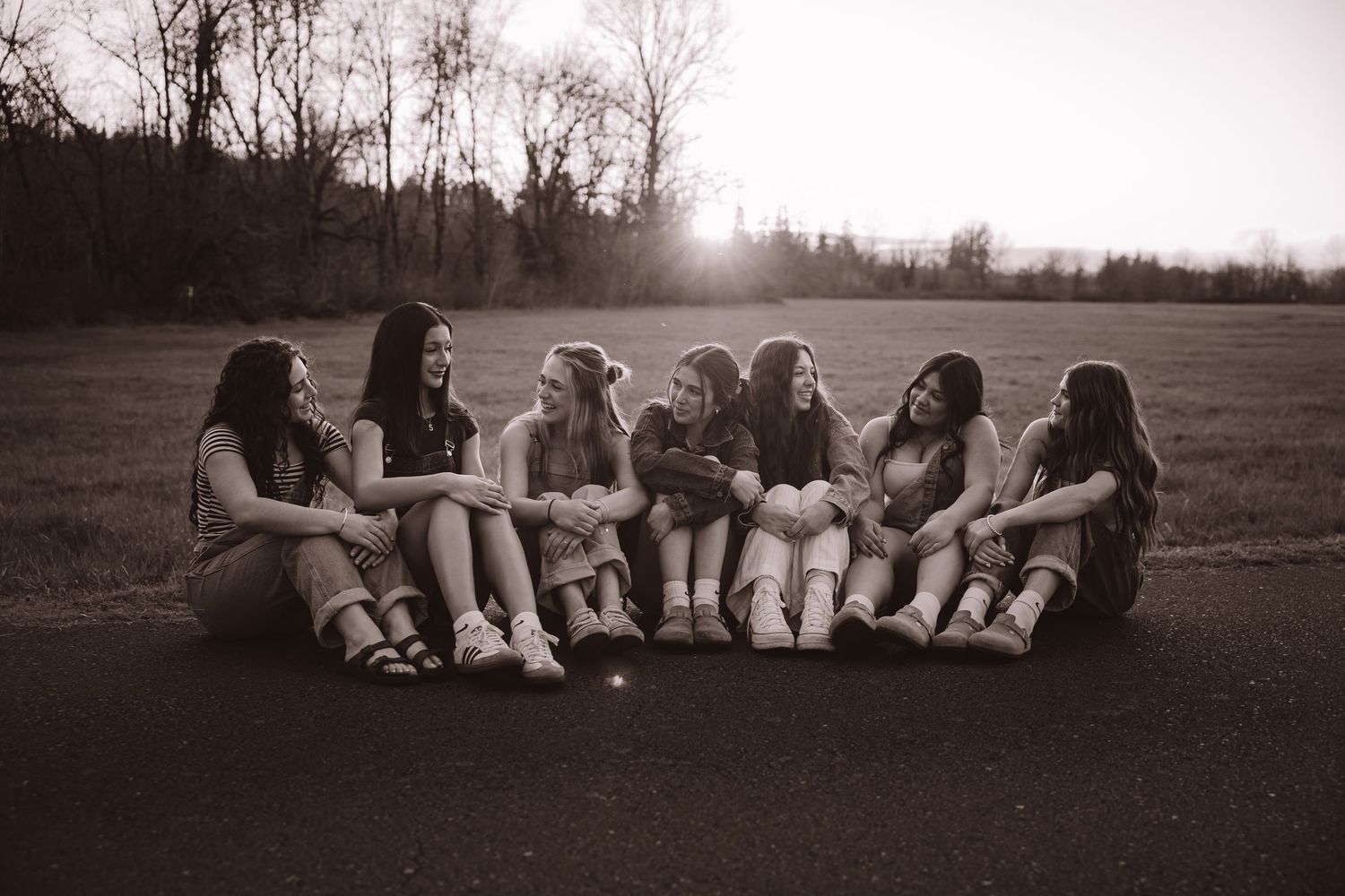Group of seven young women sitting in a circle on a road laughing in sepia tone.