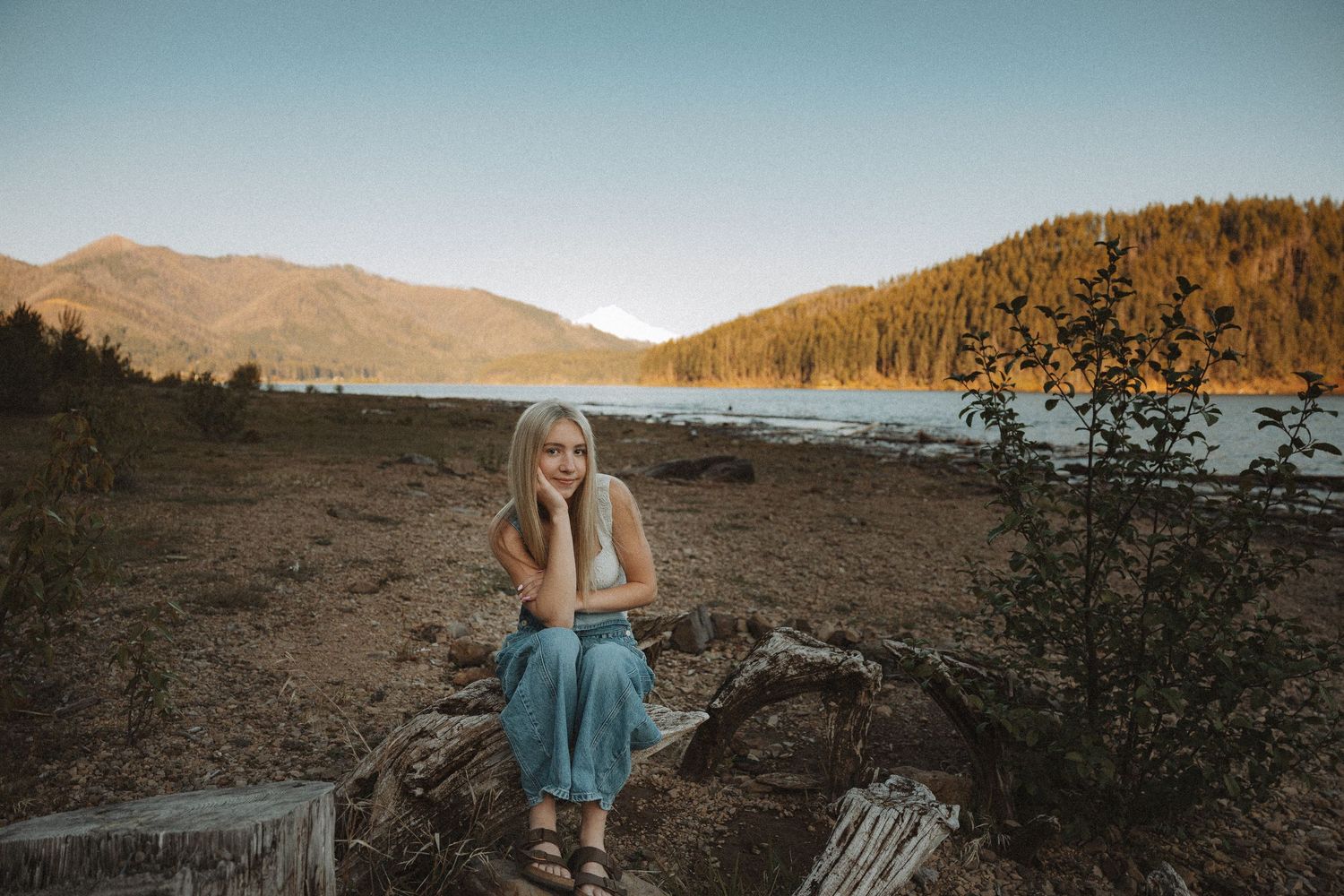 Young blonde woman sitting on driftwood by a mountain lake at sunset in a blue skirt.