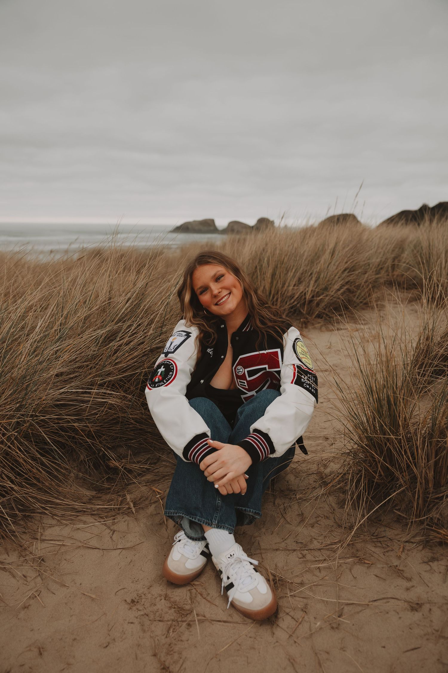 Candid portrait of someone sitting in tall beach grass wearing a varsity jacket and white sneakers.