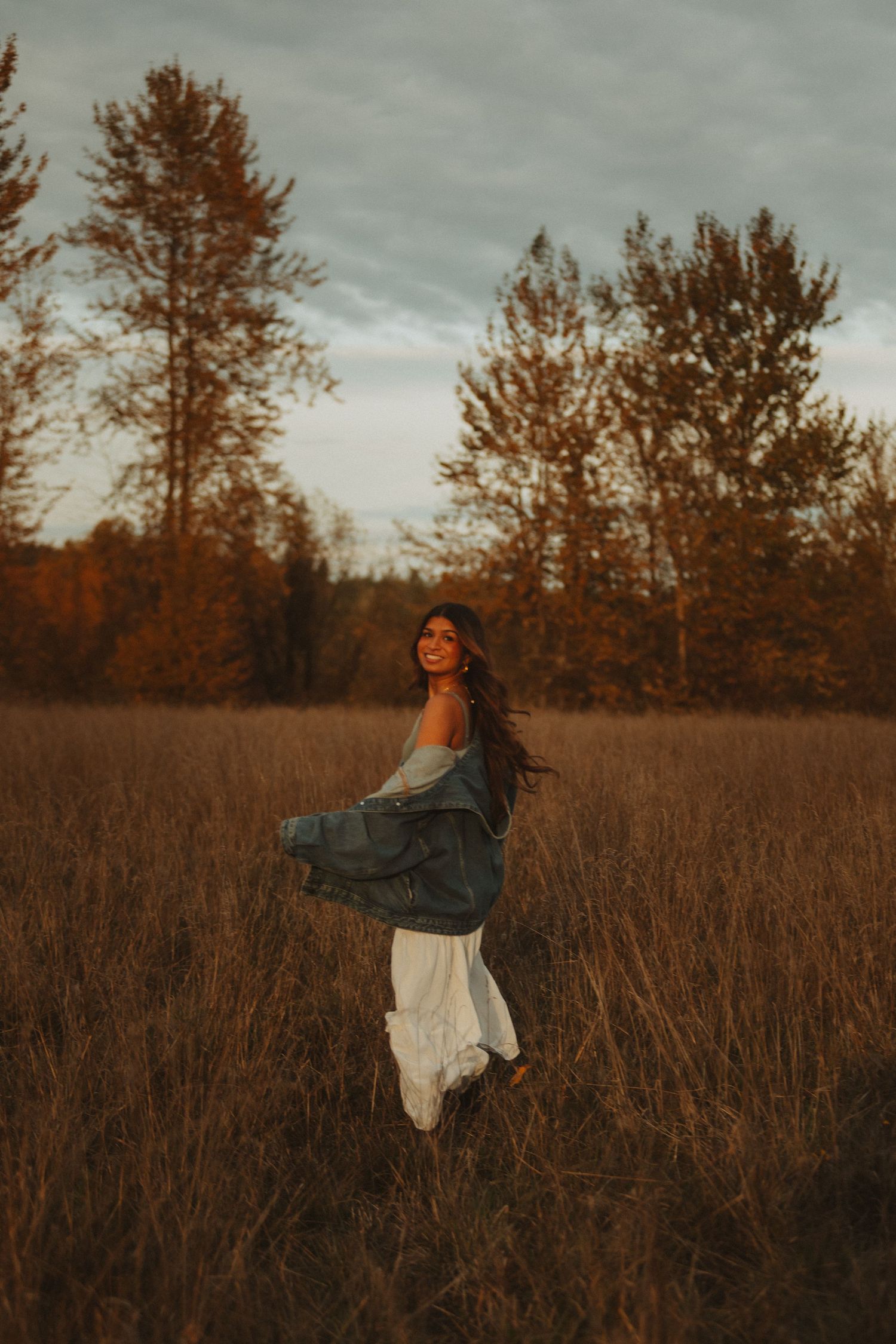 Person twirling in white dress and denim jacket through golden autumn field at dusk.