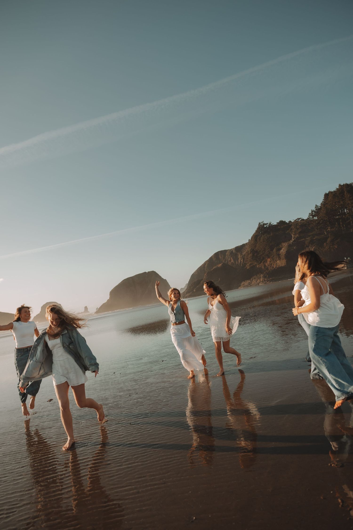 Group of friends in white dresses running joyfully on a sandy beach at golden hour with rocky cliffs in background.