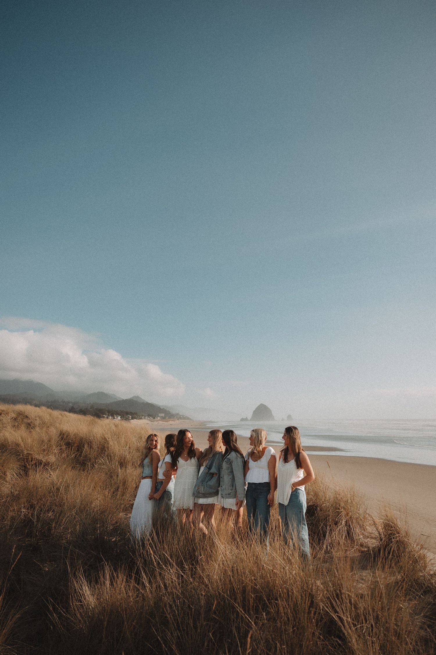 Six women gathered in golden beach grass with Haystack Rock and ocean views at Cannon Beach.