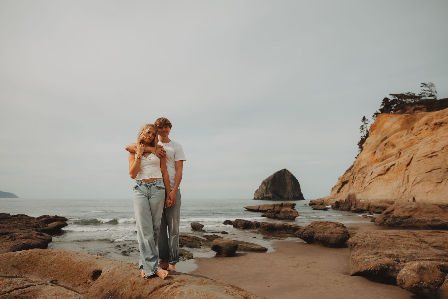 Couple embracing on a rocky beach with sea stacks and ocean waves in the background on an overcast day.