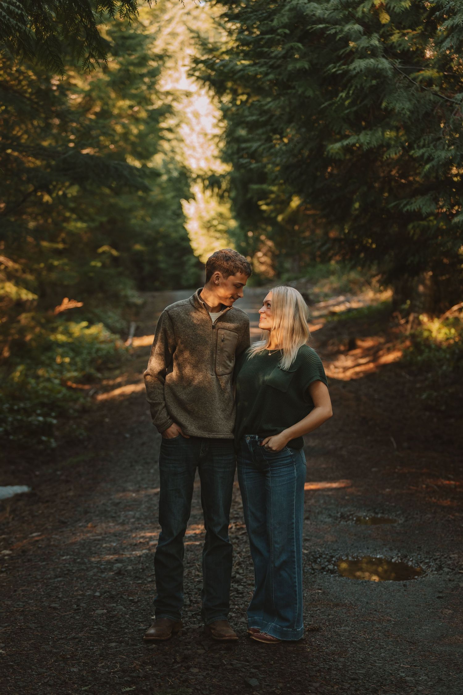 A couple shares an intimate moment on a wooded trail illuminated by golden sunlight filtering through trees.