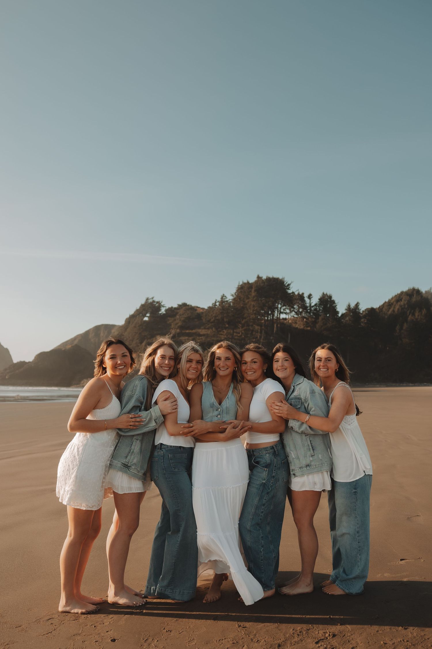 Seven women in white and denim outfits pose together on a sandy beach with ocean and trees in background.