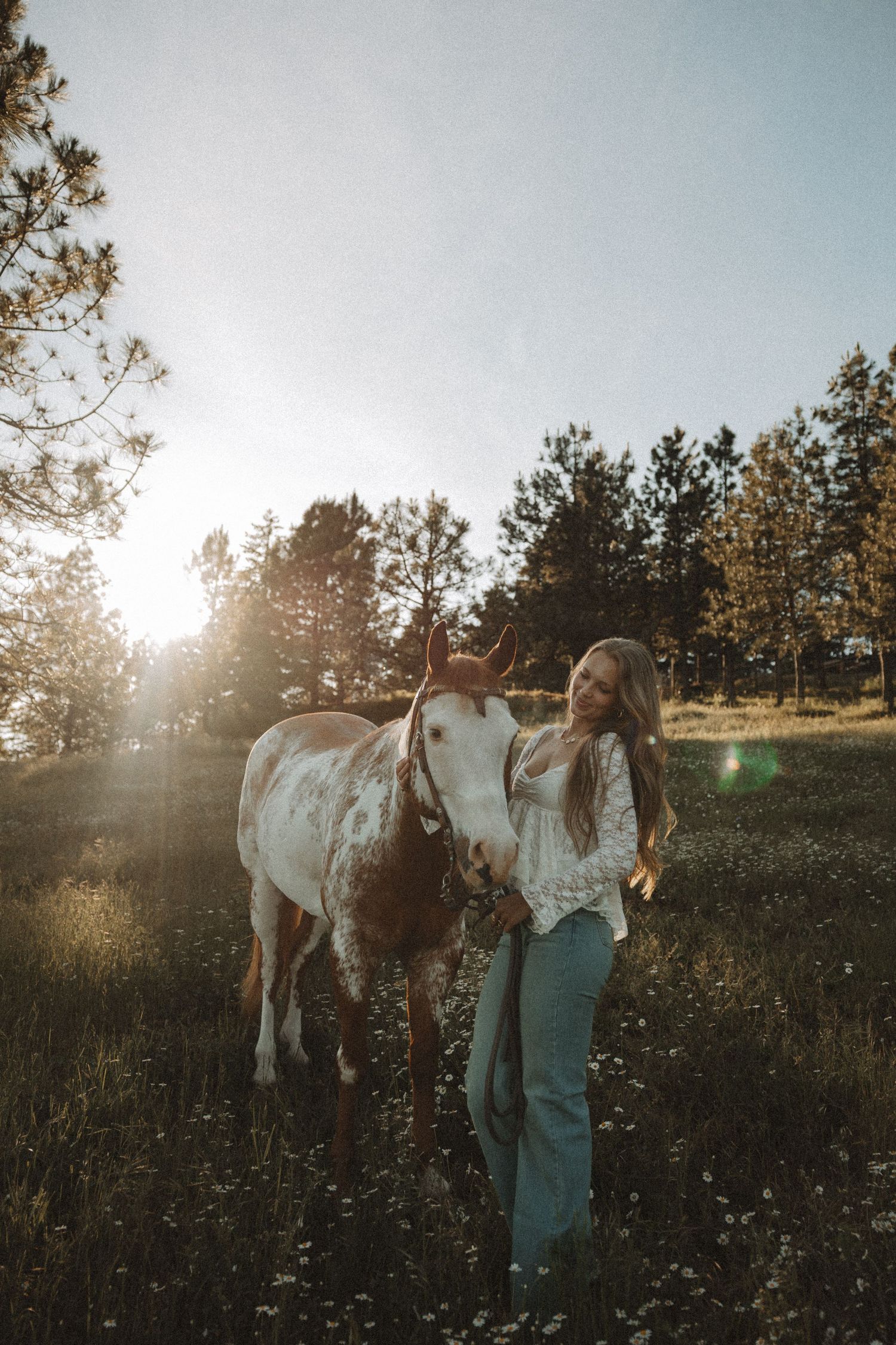 Young woman in white blouse and jeans stands beside a paint horse in a sunlit meadow with trees in background.
