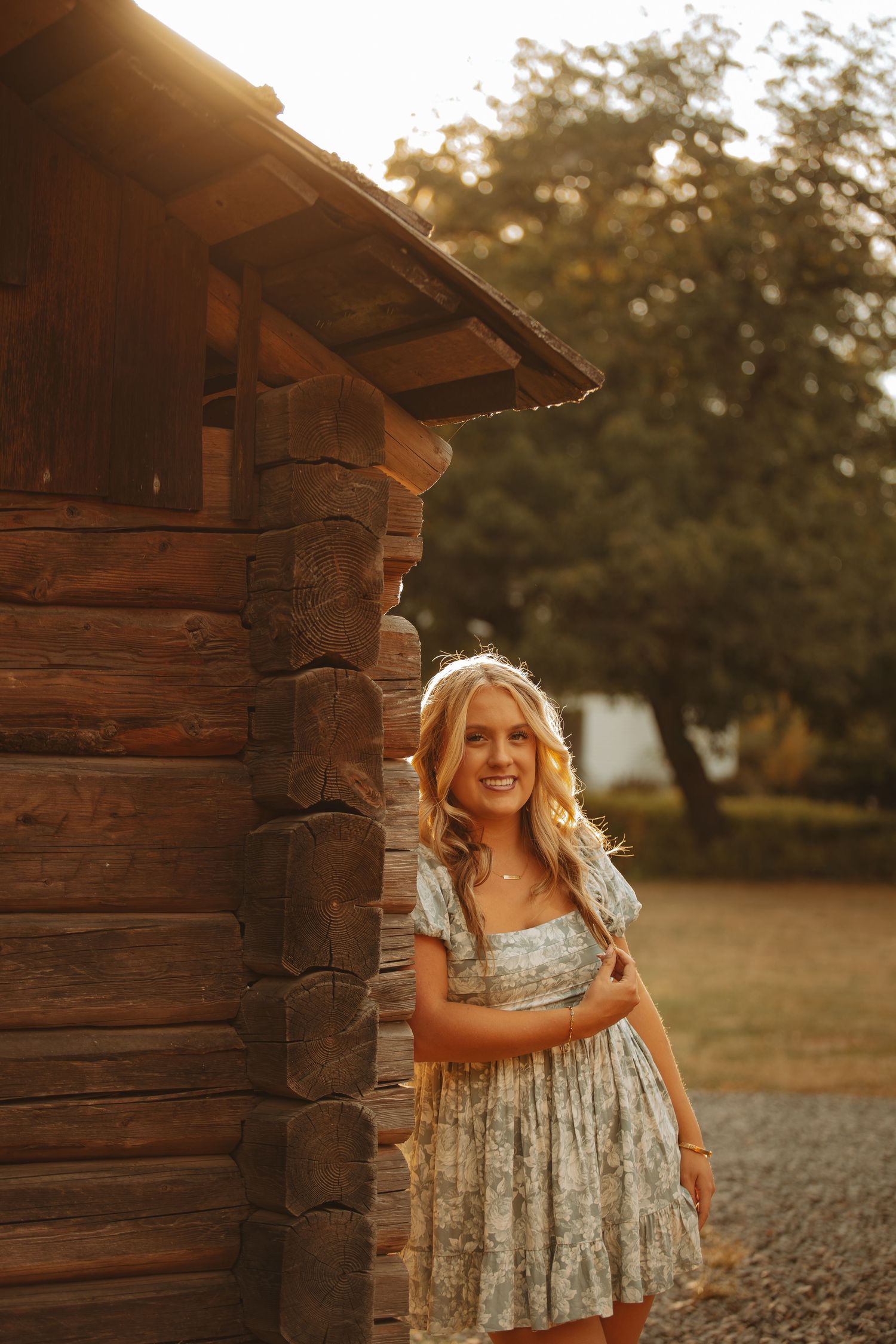 Smiling blonde woman in floral dress standing beside rustic log cabin at golden hour sunset.