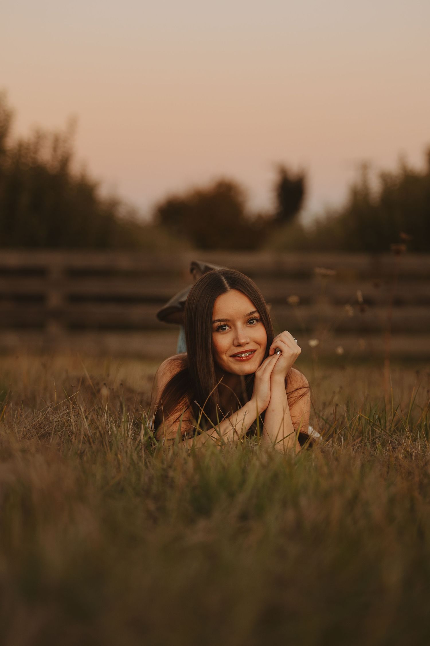Young woman lying in golden grass at sunset, smiling with hands under chin, near a wooden fence in a rural field.