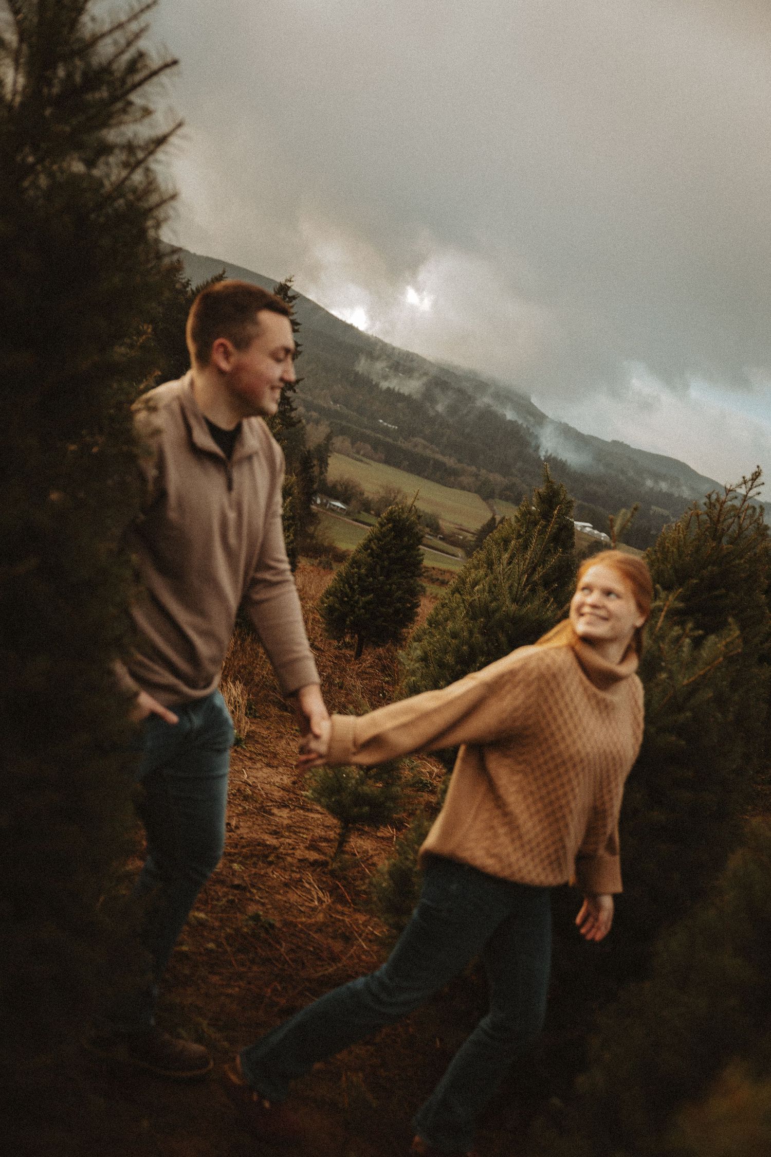 A playful couple in casual attire dances together against a scenic mountain backdrop on an overcast day.