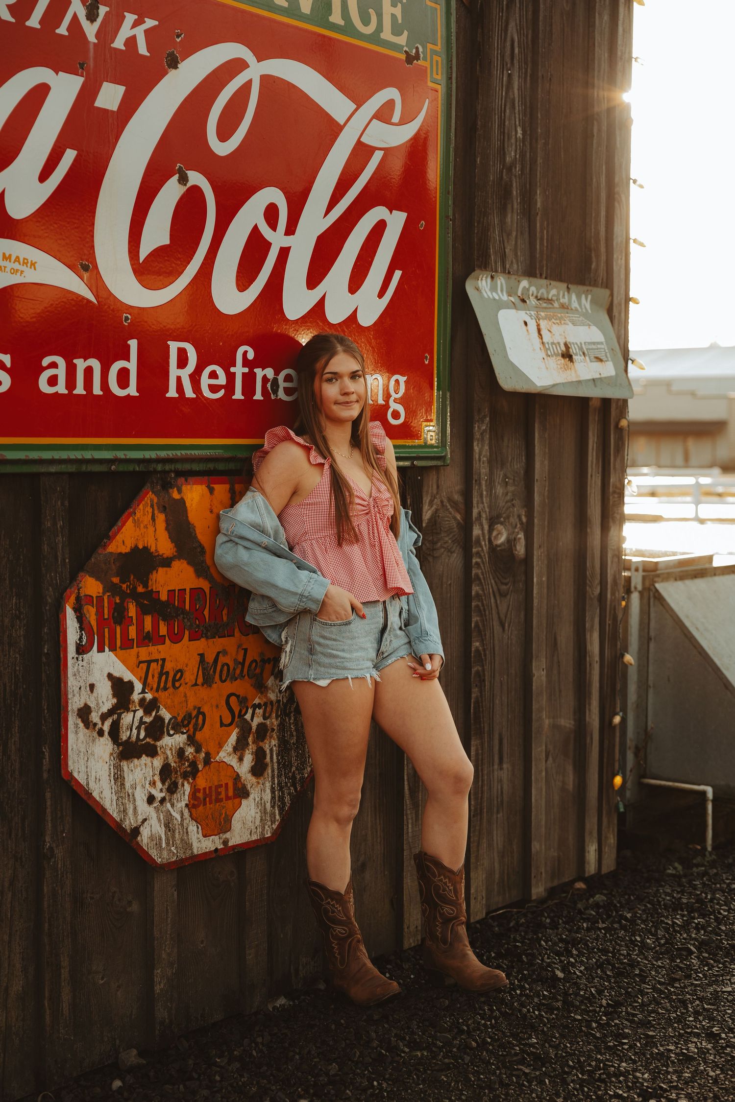 Young woman in denim jacket, pink top, jean shorts, and cowboy boots posing by vintage Coca-Cola sign at golden hour.