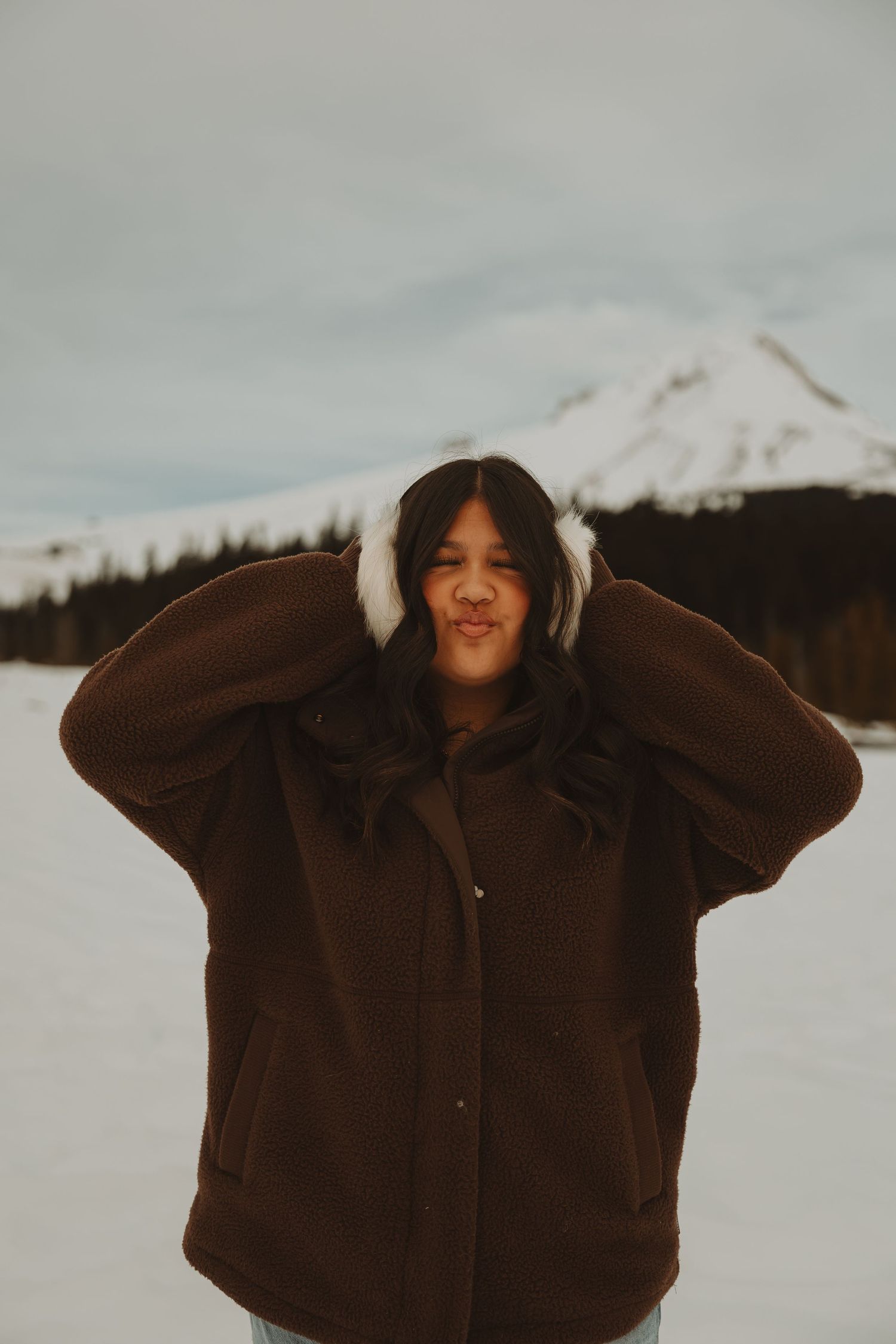 Young woman in brown coat with white earmuffs posing in snowy mountain setting.