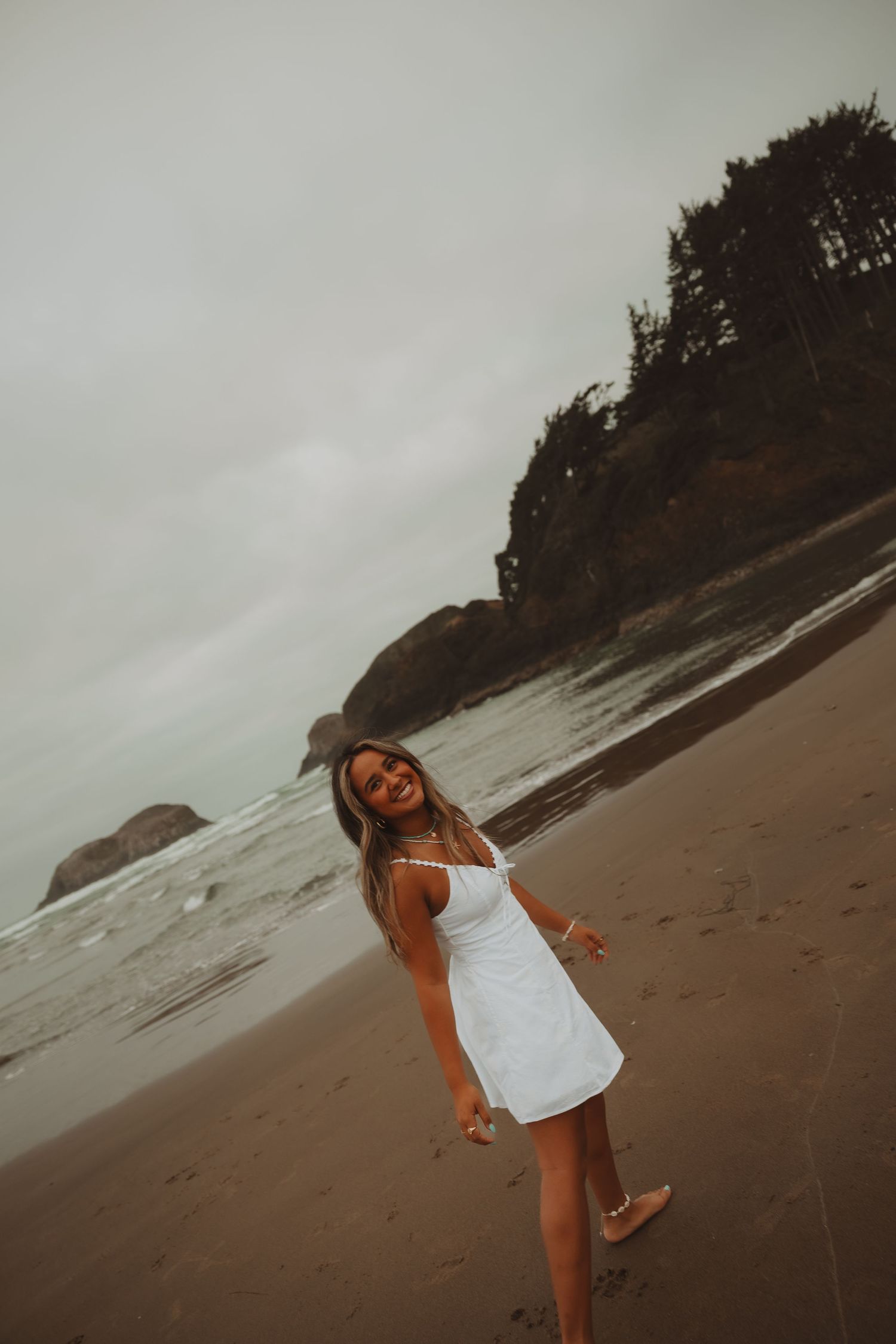 A smiling woman in a white sundress poses on a sandy beach with rocky cliffs and ocean waves in the background.
