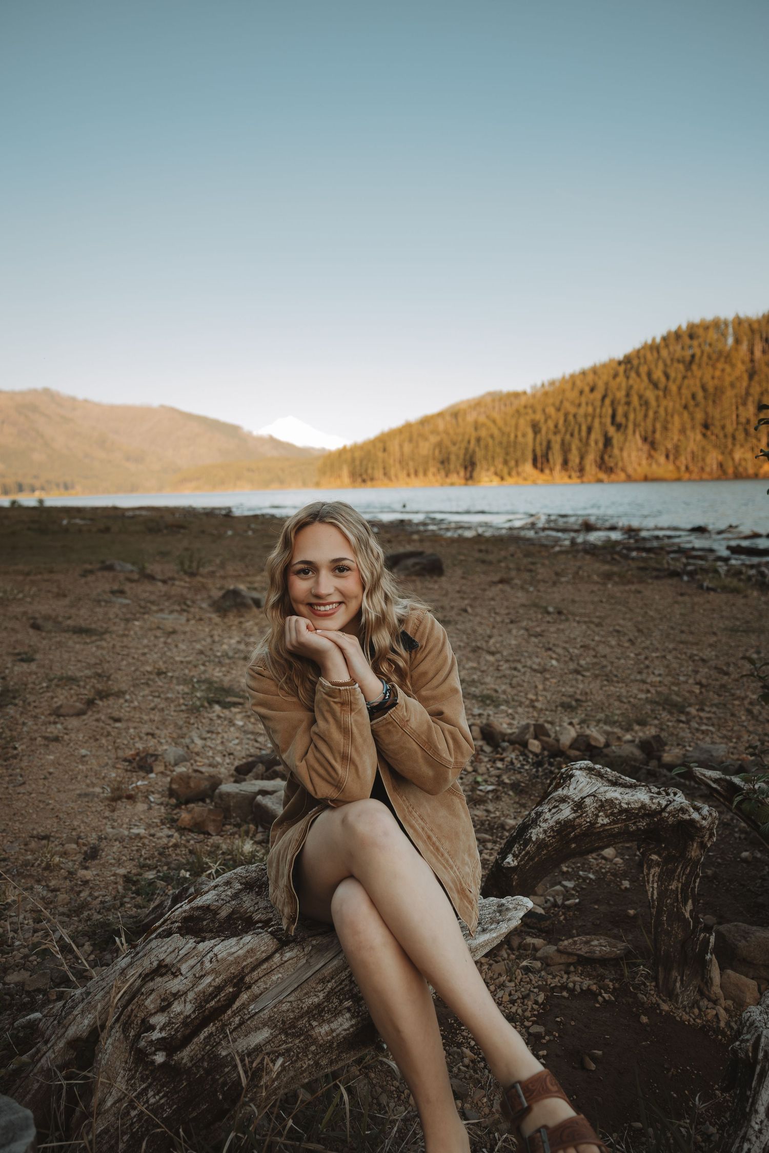 Young woman in brown jacket sitting on driftwood log by mountain lake at sunset, smiling at camera.