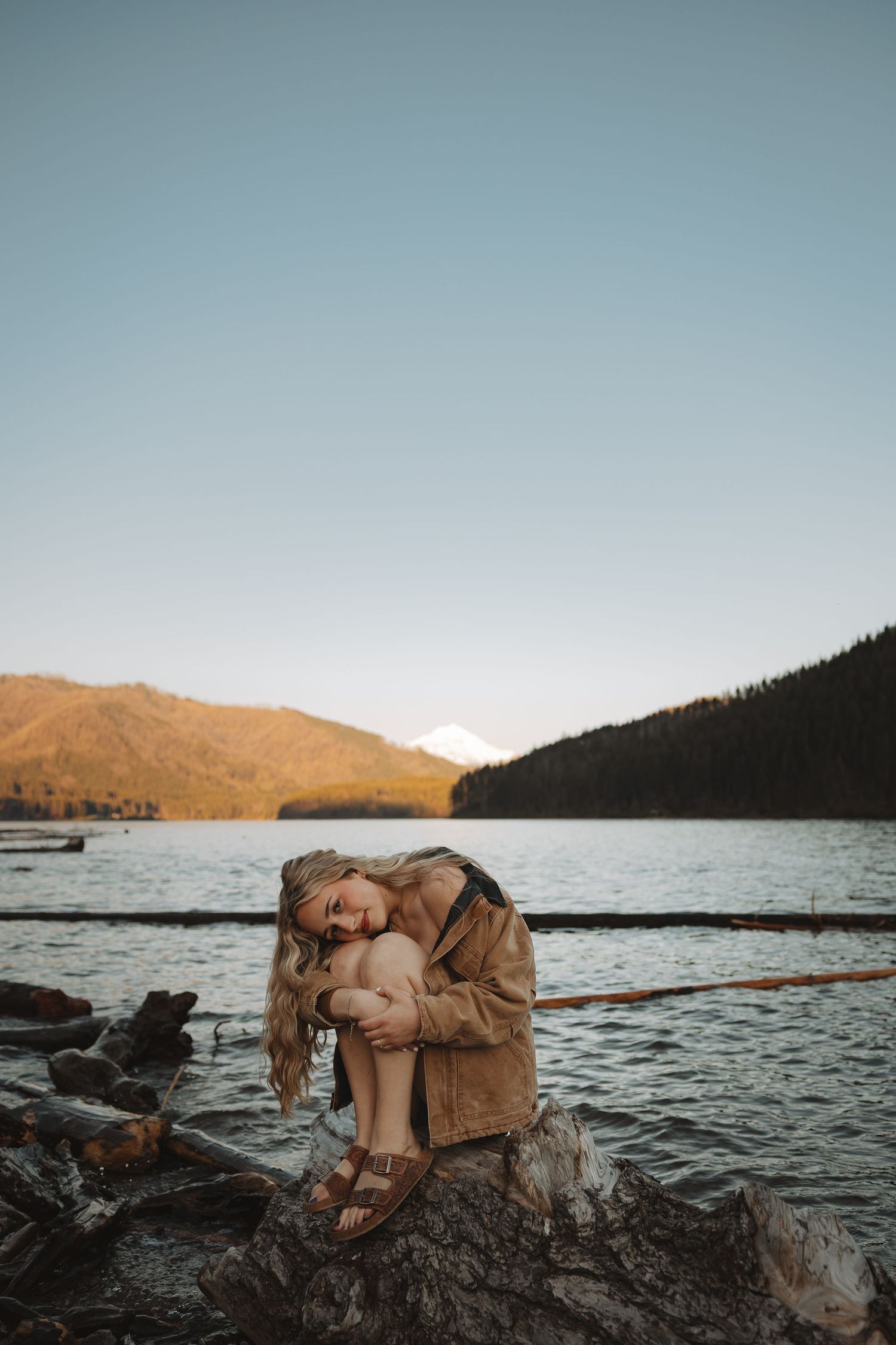 Blonde woman in tan jacket sitting on driftwood by a scenic mountain lake.