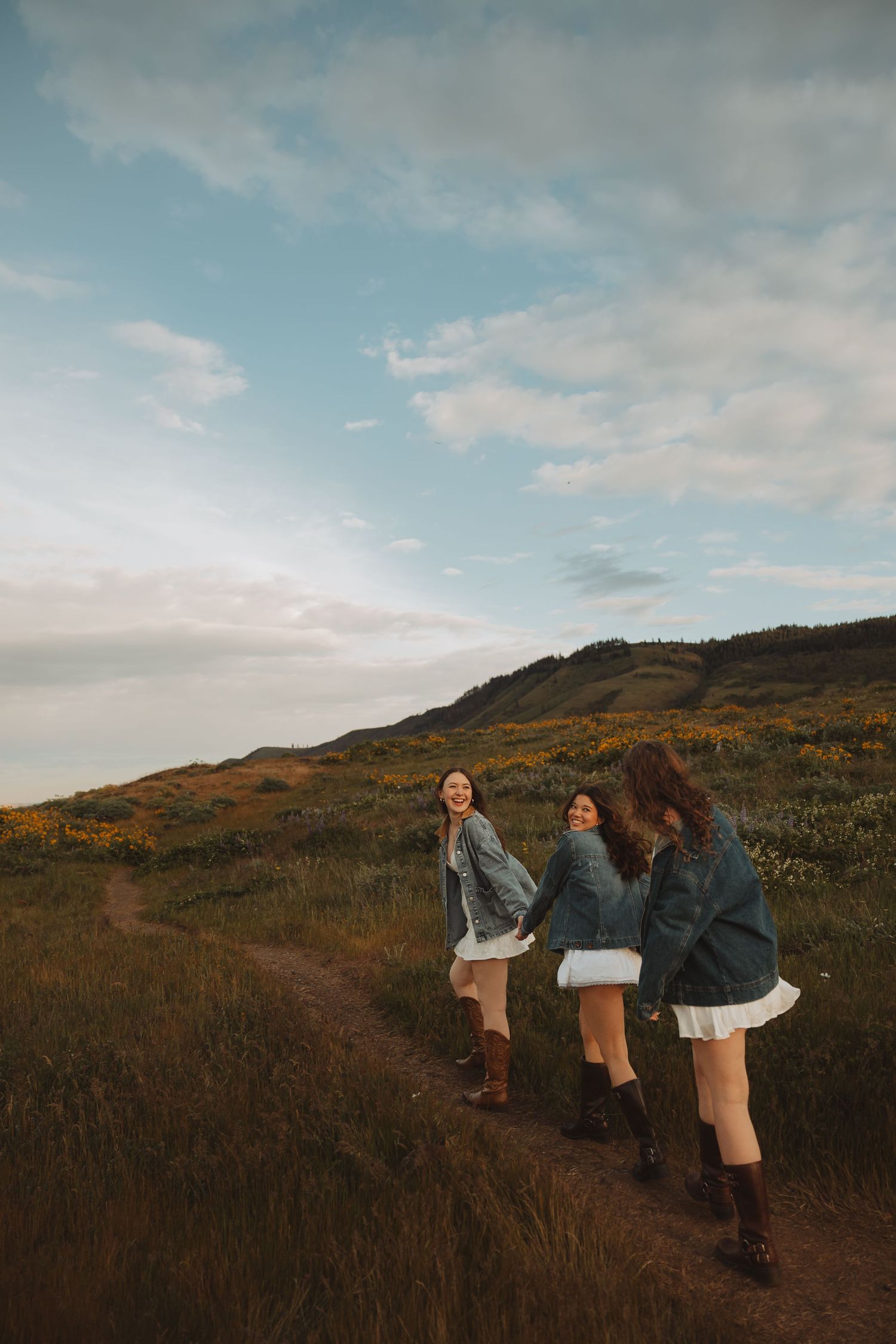Three young women in denim jackets and white skirts laugh together on a scenic hillside trail with yellow wildflowers.