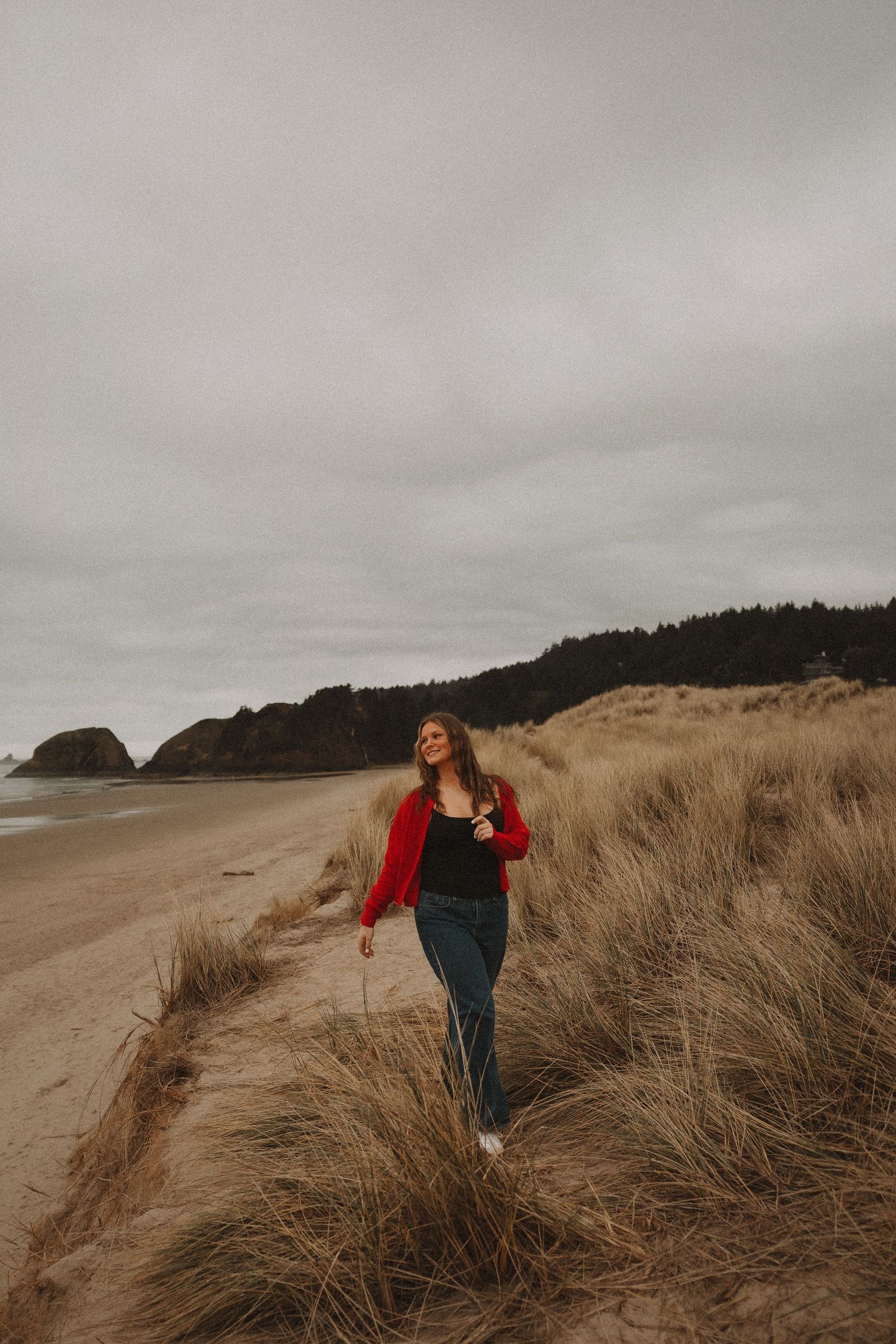 Figure in a red sweater poses playfully against moody coastal dunes with tall grass on an overcast day.