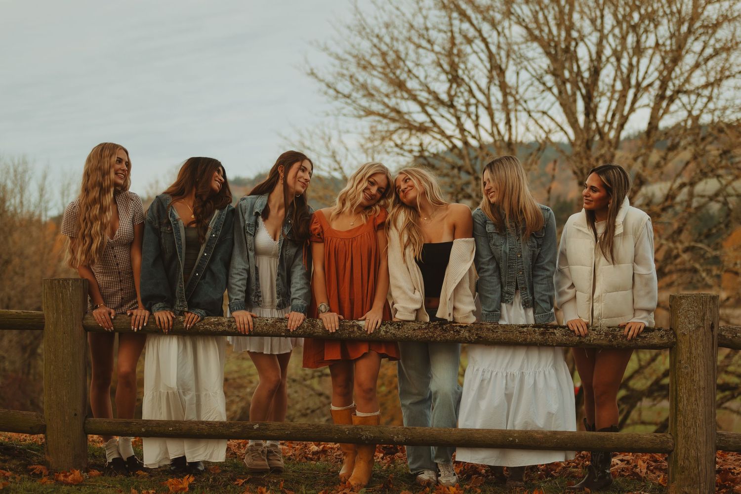 A group stands along a wooden fence in autumn, with bare trees and fallen leaves creating a rustic outdoor scene.