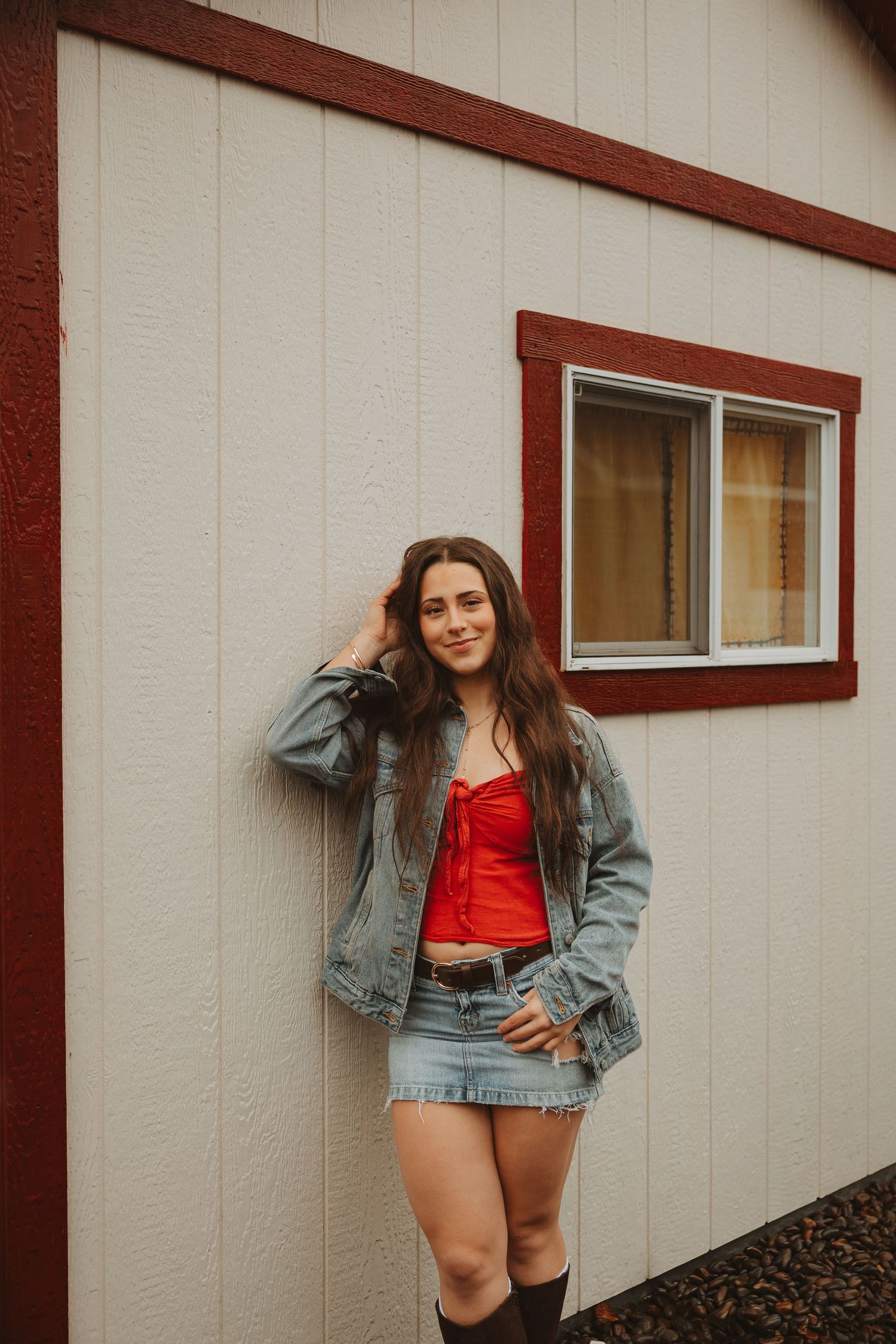 Person in denim jacket and red top poses casually against beige building wall.