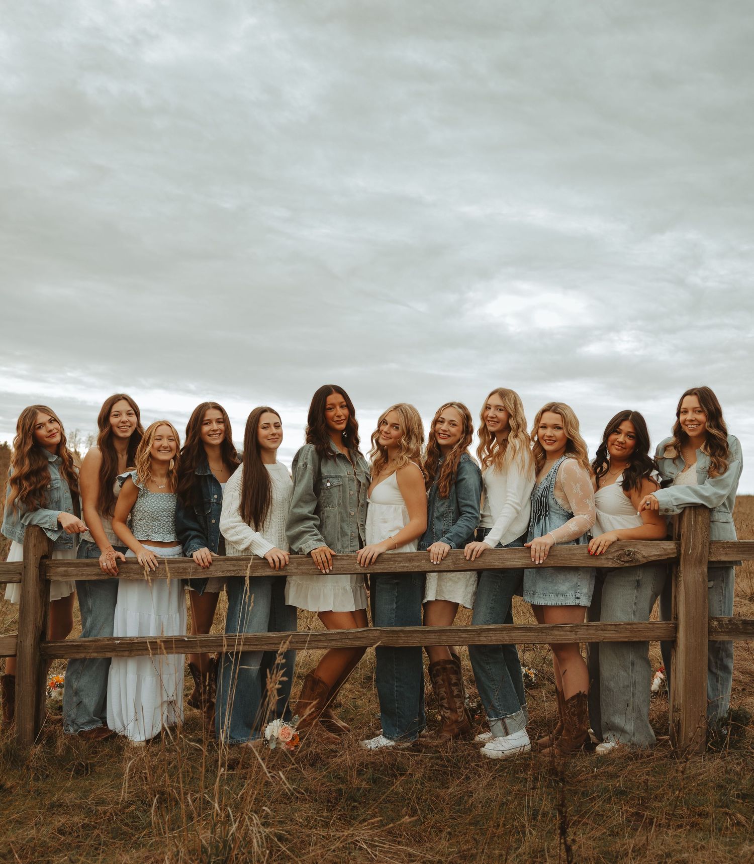 A group of friends in denim and white dresses lean against a wooden fence in a rustic outdoor setting.