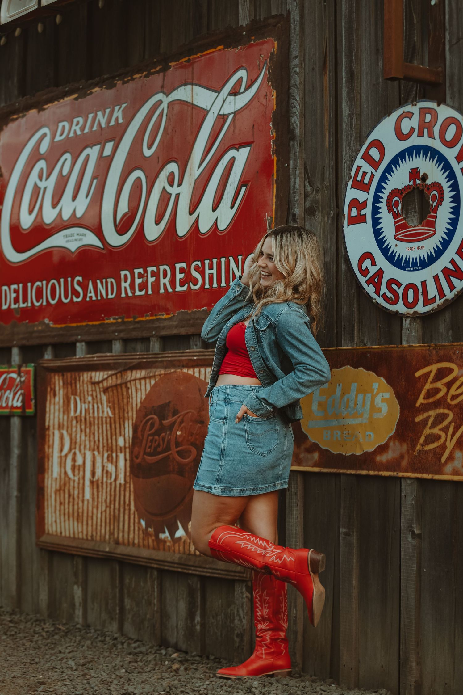 Red boots and denim outfit against rustic wall with vintage Coca-Cola and Red Crown Gasoline signs.