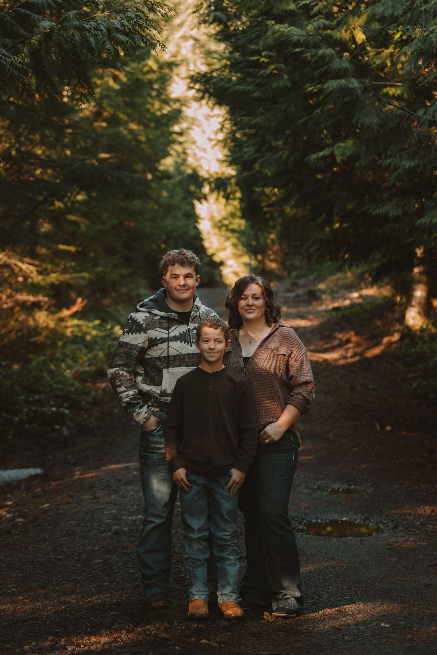 Three people pose together on a dirt path surrounded by tall evergreen trees in warm autumn lighting.