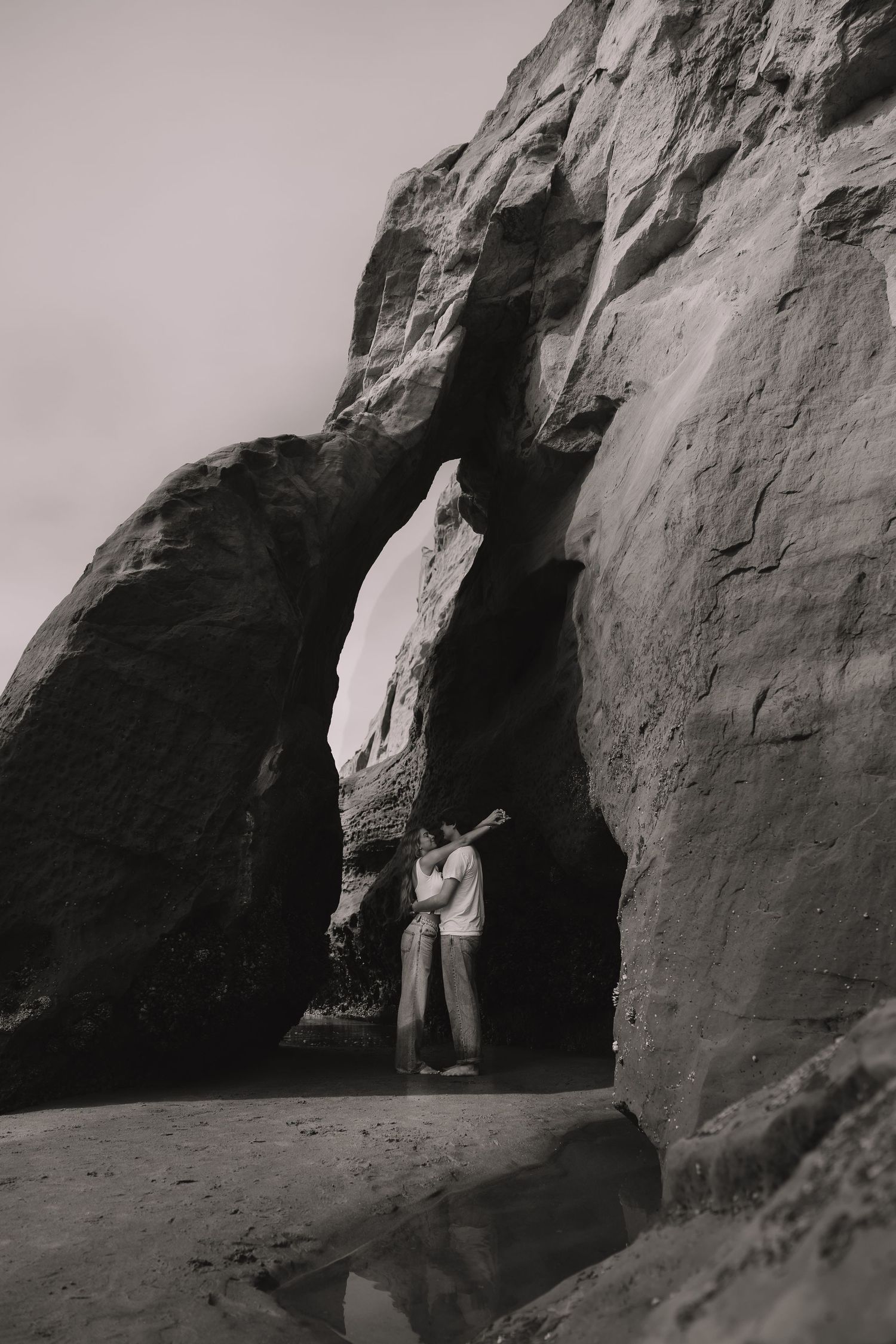 Couple embracing under a natural rock arch on a sandy beach in a romantic black and white photo.